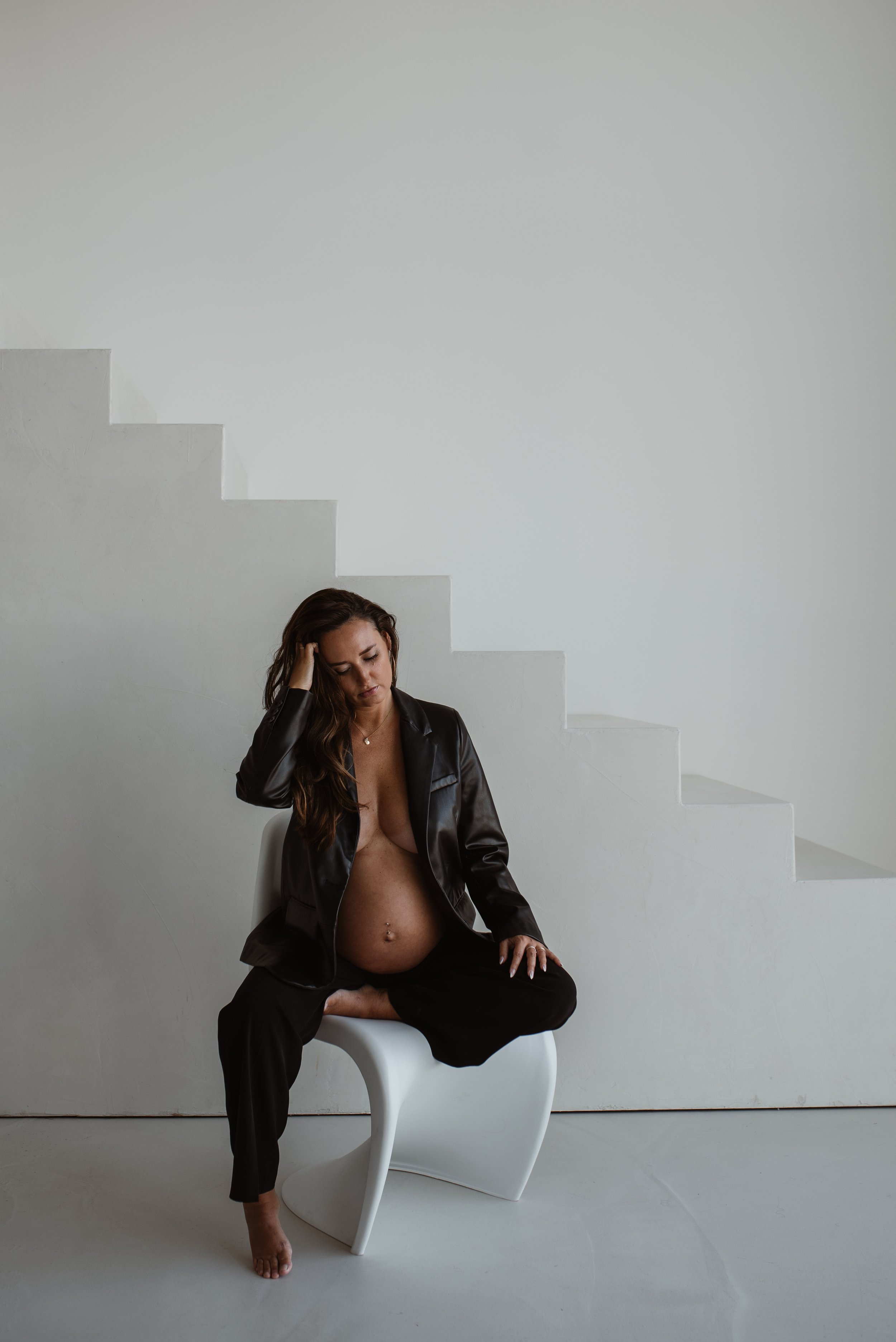 Eine Frau sitzt auf einem weißen Stuhl vor einer weißen Wand, trägt eine schwarze Lederjacke, ist unbedeckt vom Bauch bis zu den Beinen, hat braune lange Haare und scheint nach unten zu blicken, während sie mit einer Hand ihr Haar berührt.