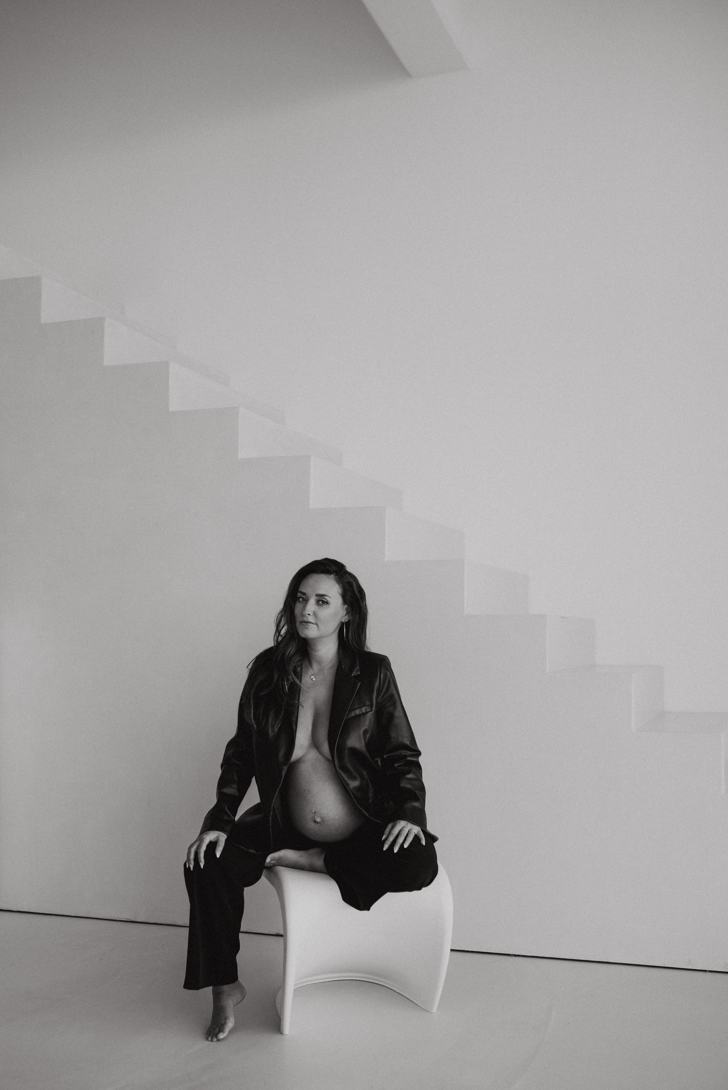 Schwarz-Weiß-Fotografie einer jungen Frau mit dunklen, lockigen Haaren, sitzend in einem schwarzen Outfit, auf einem weißen Stuhl vor einer hellen Wand mit abstrakten Stufenmustern.