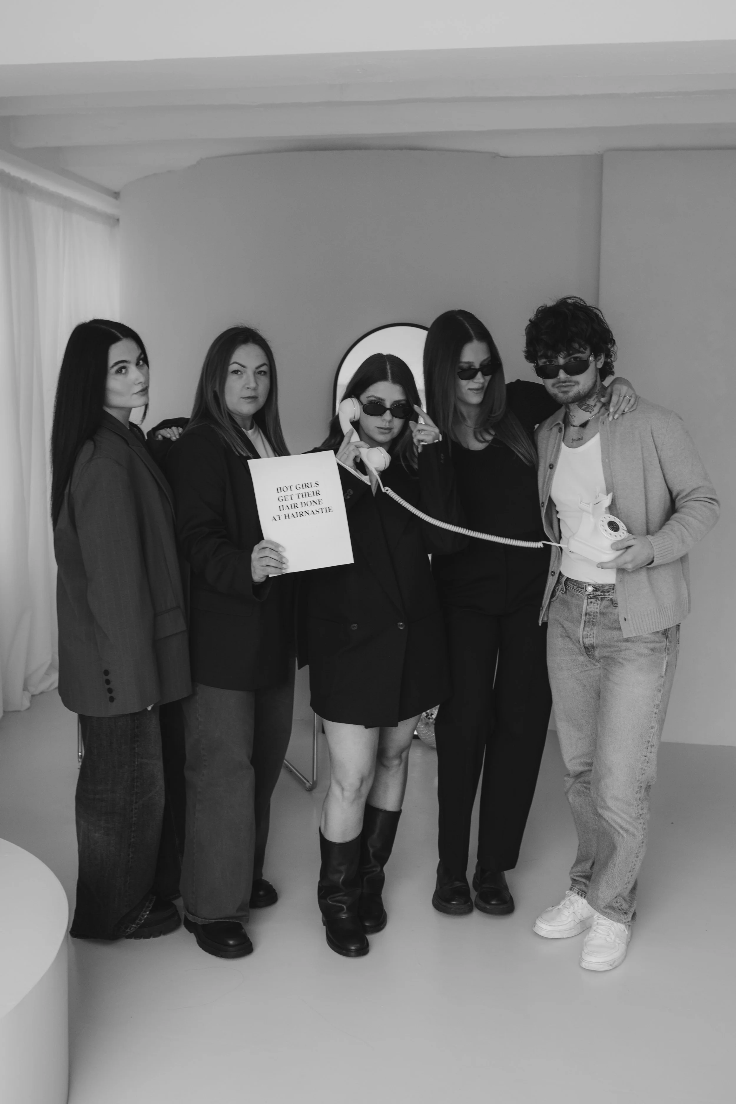 Vier Frauen stehen nebeneinander vor einem Spiegel, eine hält ein Telefon, eine trägt Sonnenbrillen, zwei schauen in die Kamera, eine hält eine Pappschild mit dem Text 'Hot Girls get their hair done at Hairnstie'. Ein Mann steht rechts, trägt eine So