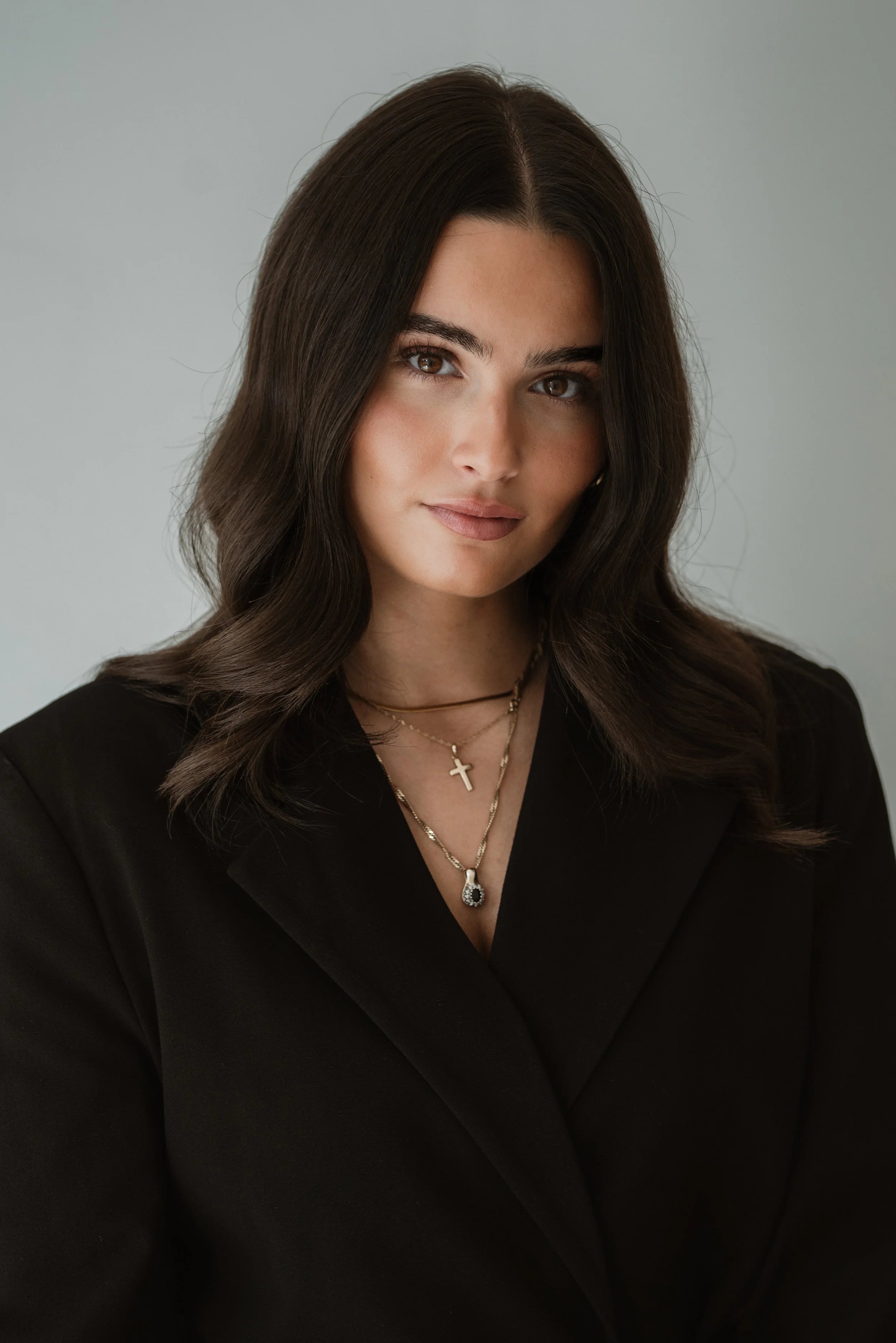 Portrait einer jungen Frau mit langem dunklem Haar, braunen Augen, in schwarzem Kleid und mehreren Halsketten, vor hellem Hintergrund.