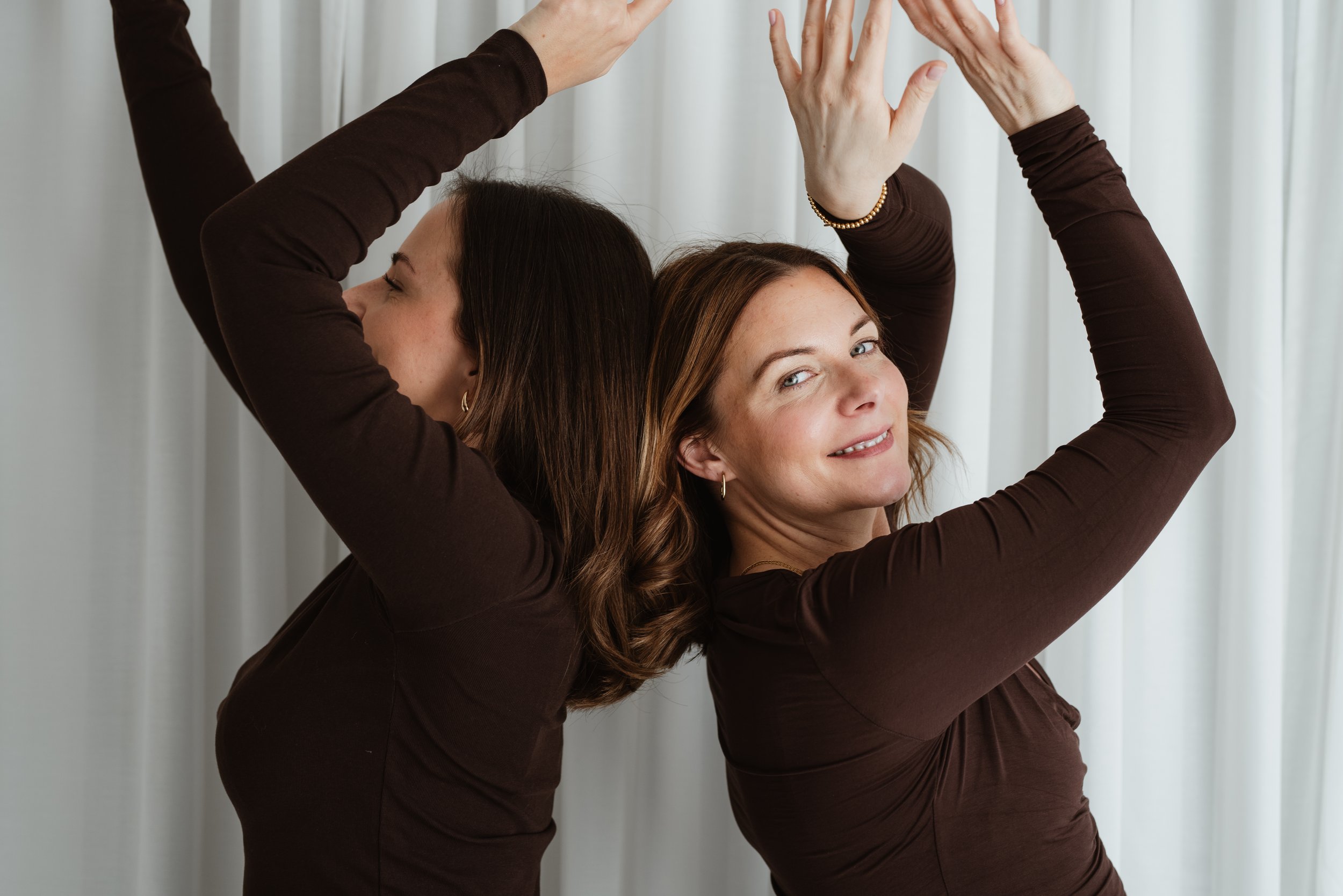 Zwei Frauen machen Yogaübungen mit ihren Armen über dem Kopf, vor einem hellen Hintergrund.