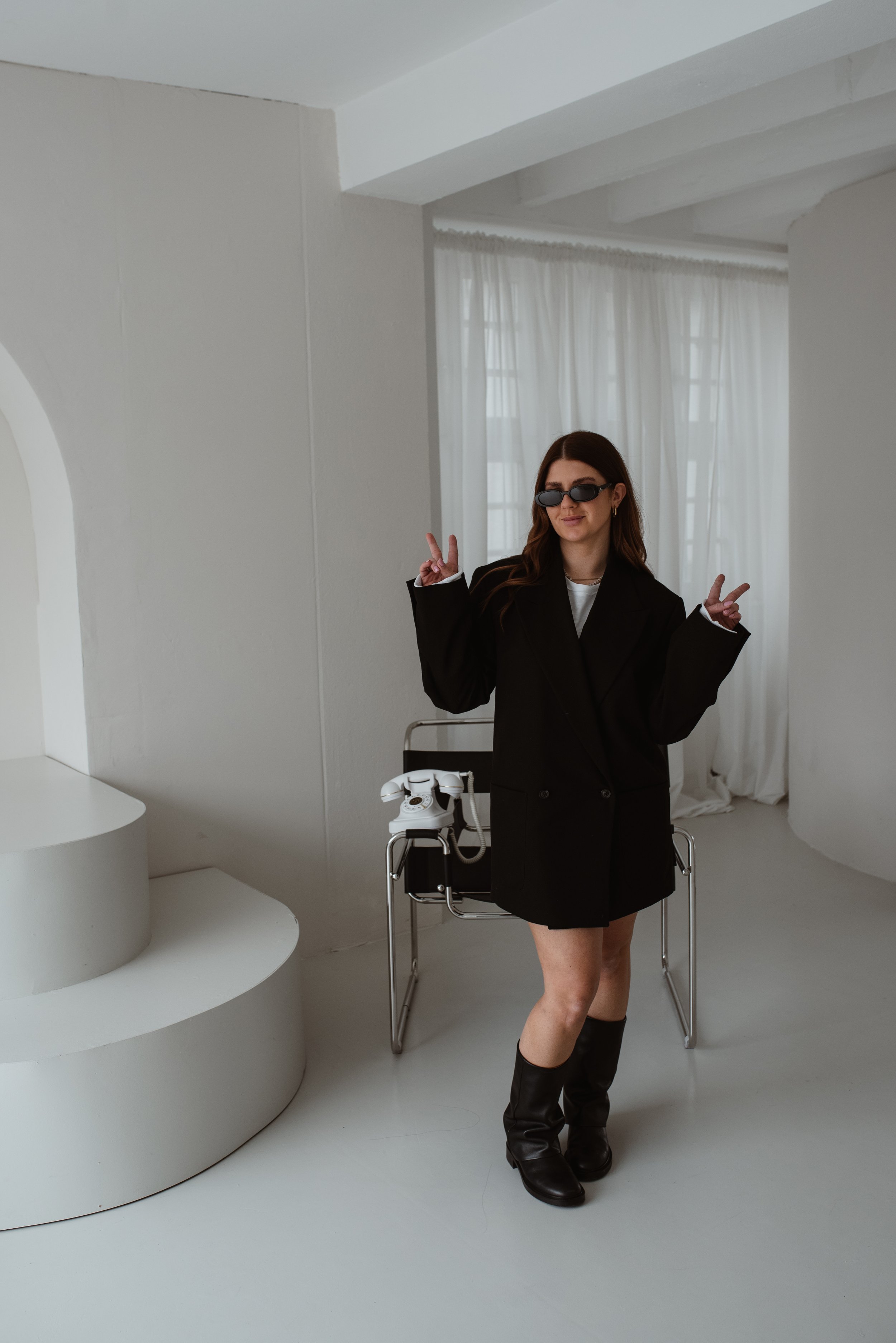 Junge Frau mit dunklen Haaren und Sonnenbrille posiert in einem weißen Raum, trägt einen schwarzen Mantel, Stiefel und zeigt Peace-Zeichen mit beiden Händen. Neben ihr steht ein Stuhl mit einer weißen Tischlampe und einem Telefondrehstuhl.