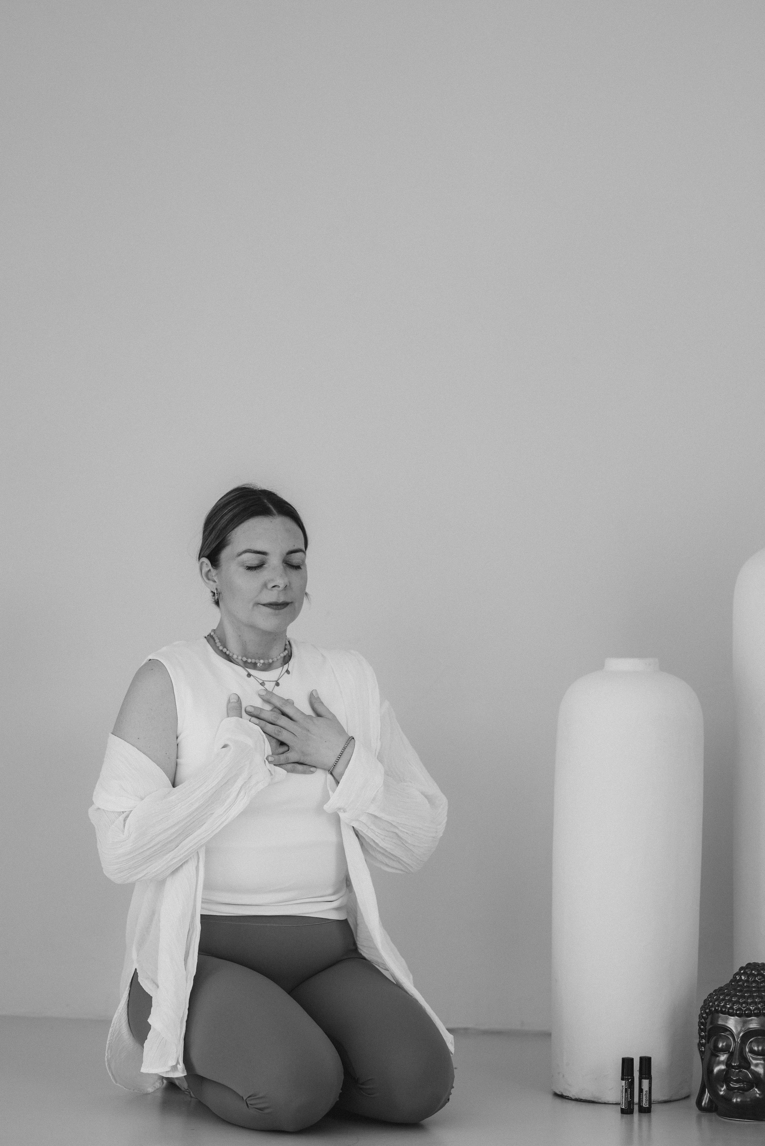 Eine Frau sitzt auf dem Boden in einer ruhigen Haltung, die Hände auf der Brust, in einem weißen Oberteil und grauen Hosen, vor einem weißen Wand mit einer weißen Vase, einem schwarzen Buddha-Kopf und einigen schwarzen Stiften.