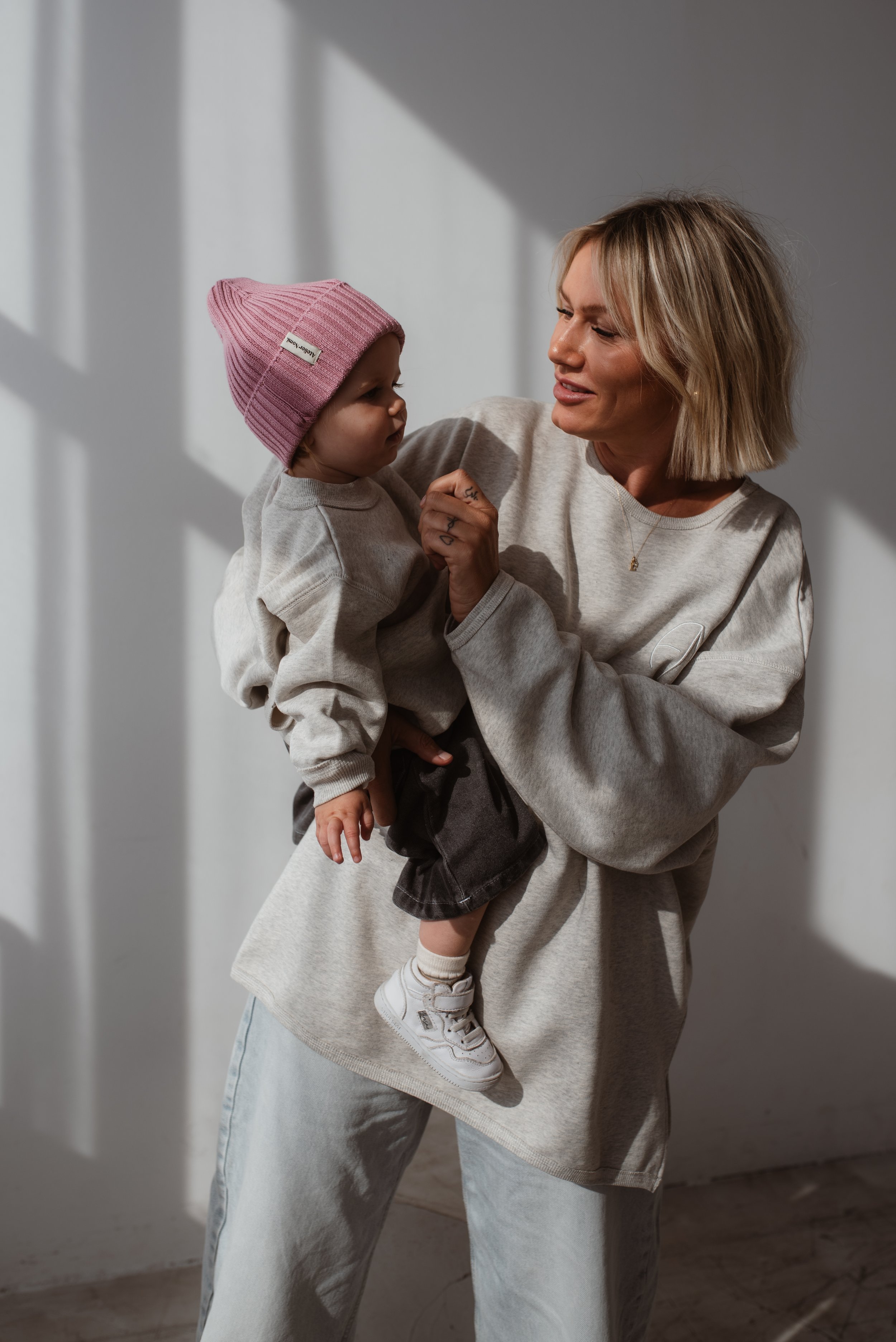 Eine Frau hält ein Kind in den Armen, beide schauen sich an. Das Kind trägt eine rosa Mütze und ein graues Sweatshirt, die Frau trägt ein beige Sweatshirt und weite Hosen. Das Bild ist in einem Raum mit natürlichem Licht, das durch Fenster fällt.