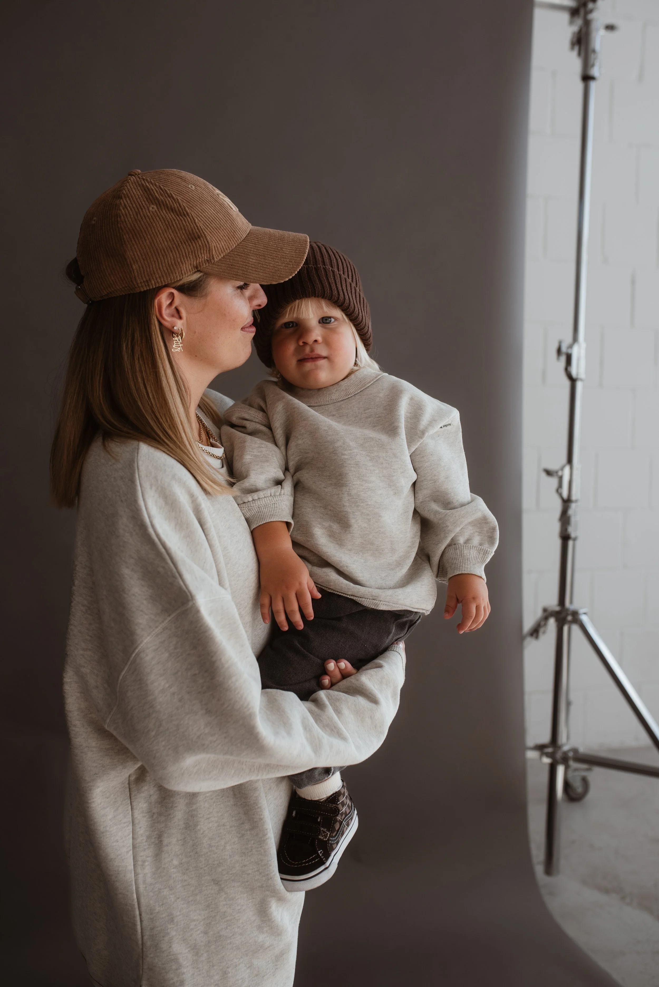Eine Frau hält ein kleines Kind in den Armen. Beide tragen beige bzw. braune Kleidung und Mützen. Sie stehen vor einem grauen Hintergrund in einem Fotostudio.