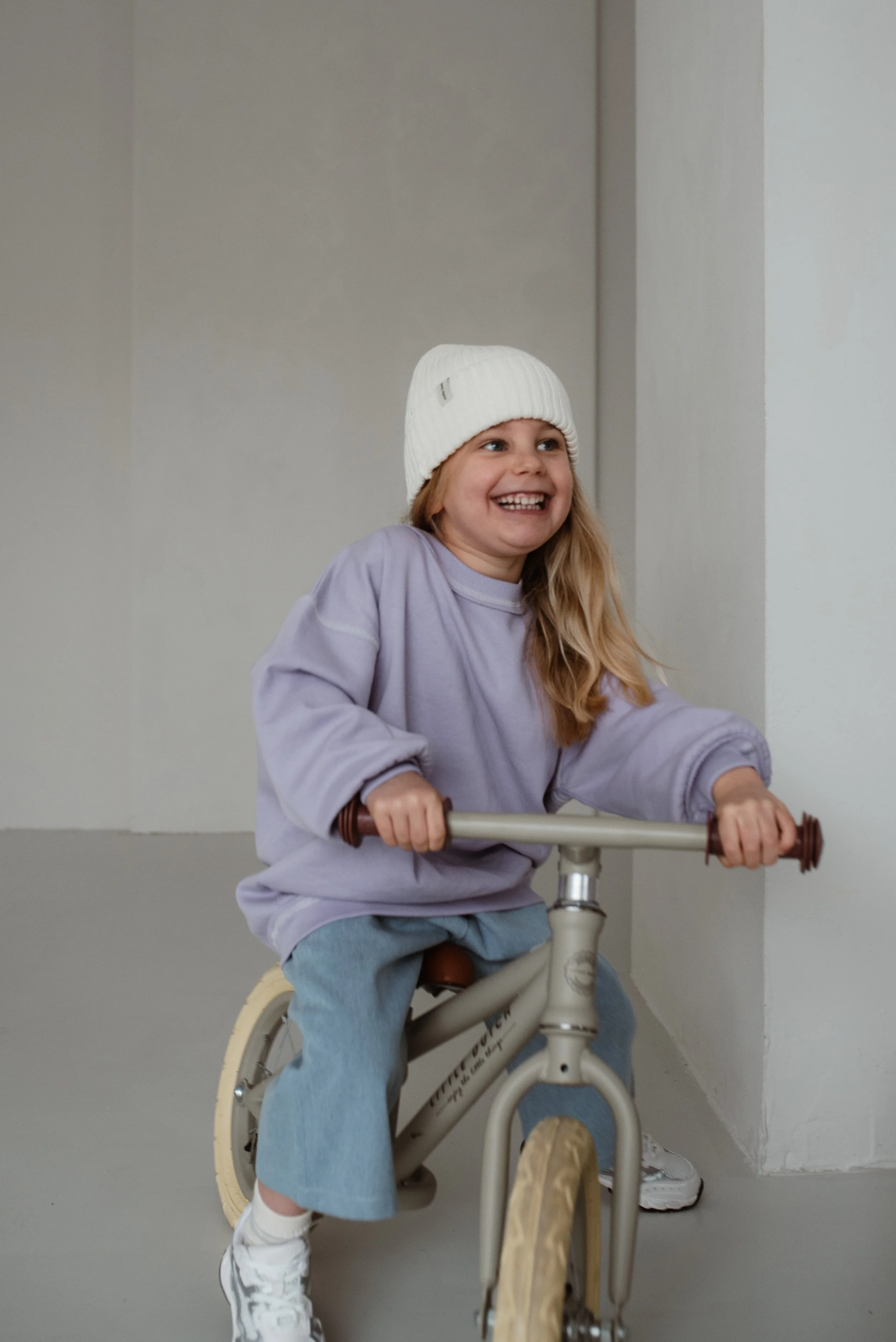 Ein lachendes Mädchen mit einem weißen Wintermütze fährt auf einem Fahrrad in einem Raum mit weißen Wänden.