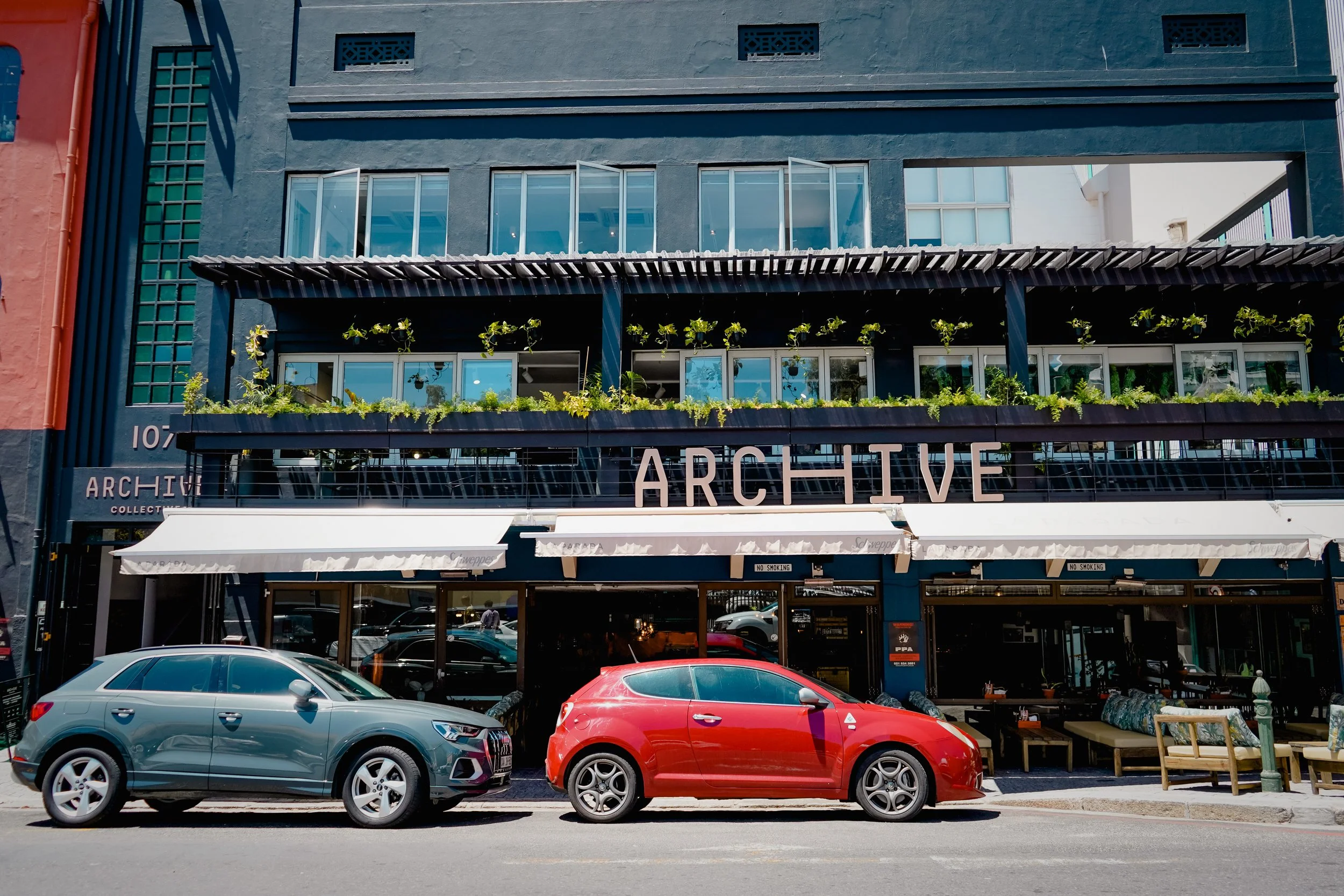 Facade of a multi-story building with storefront, including a sign that reads 'ARCHIVE' and some greenery on a balcony, with parked cars in front.