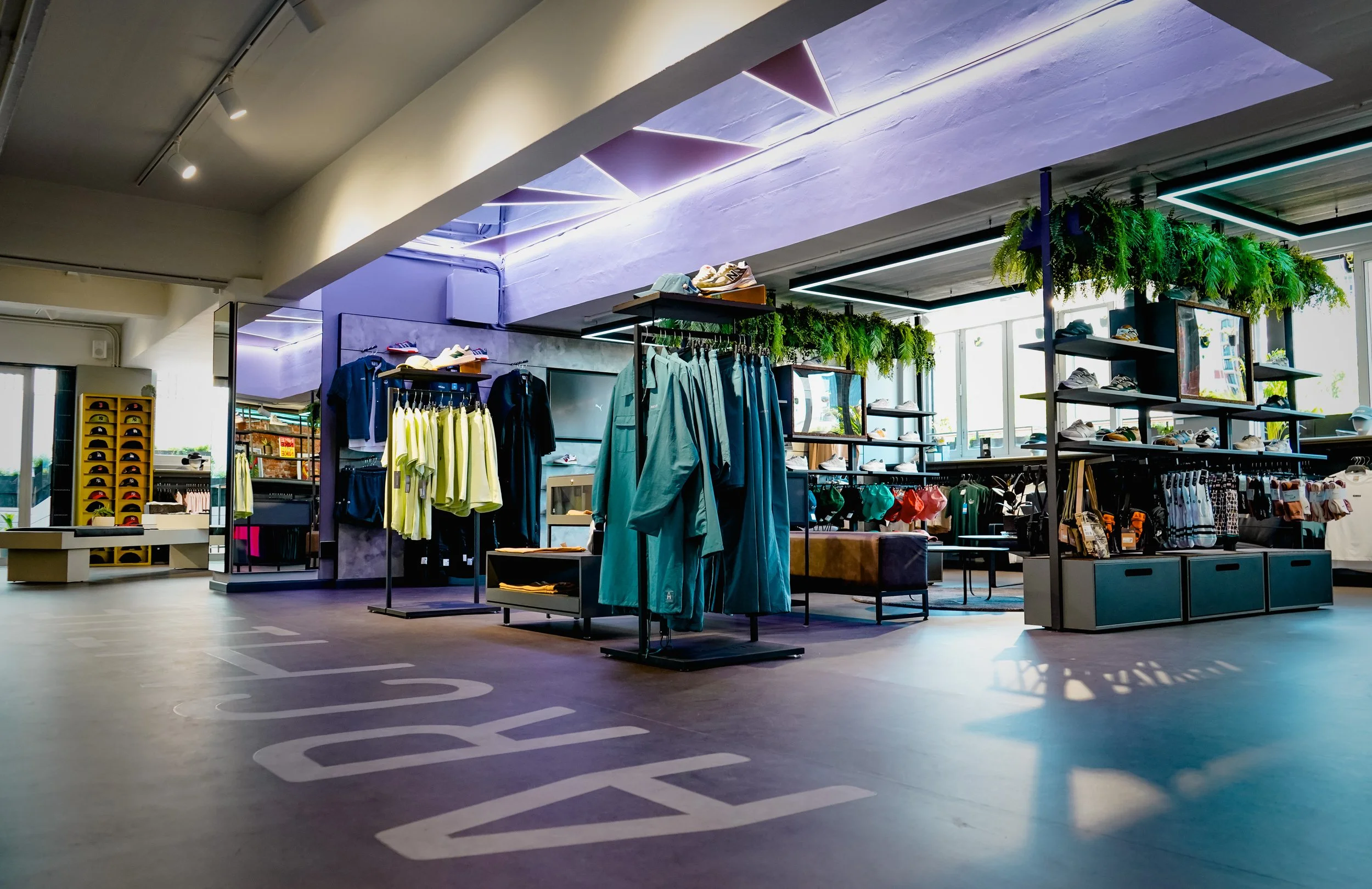 Indoor clothing store with racks of garments, shoes on shelves, and hanging plants near large windows, under a skylight ceiling.