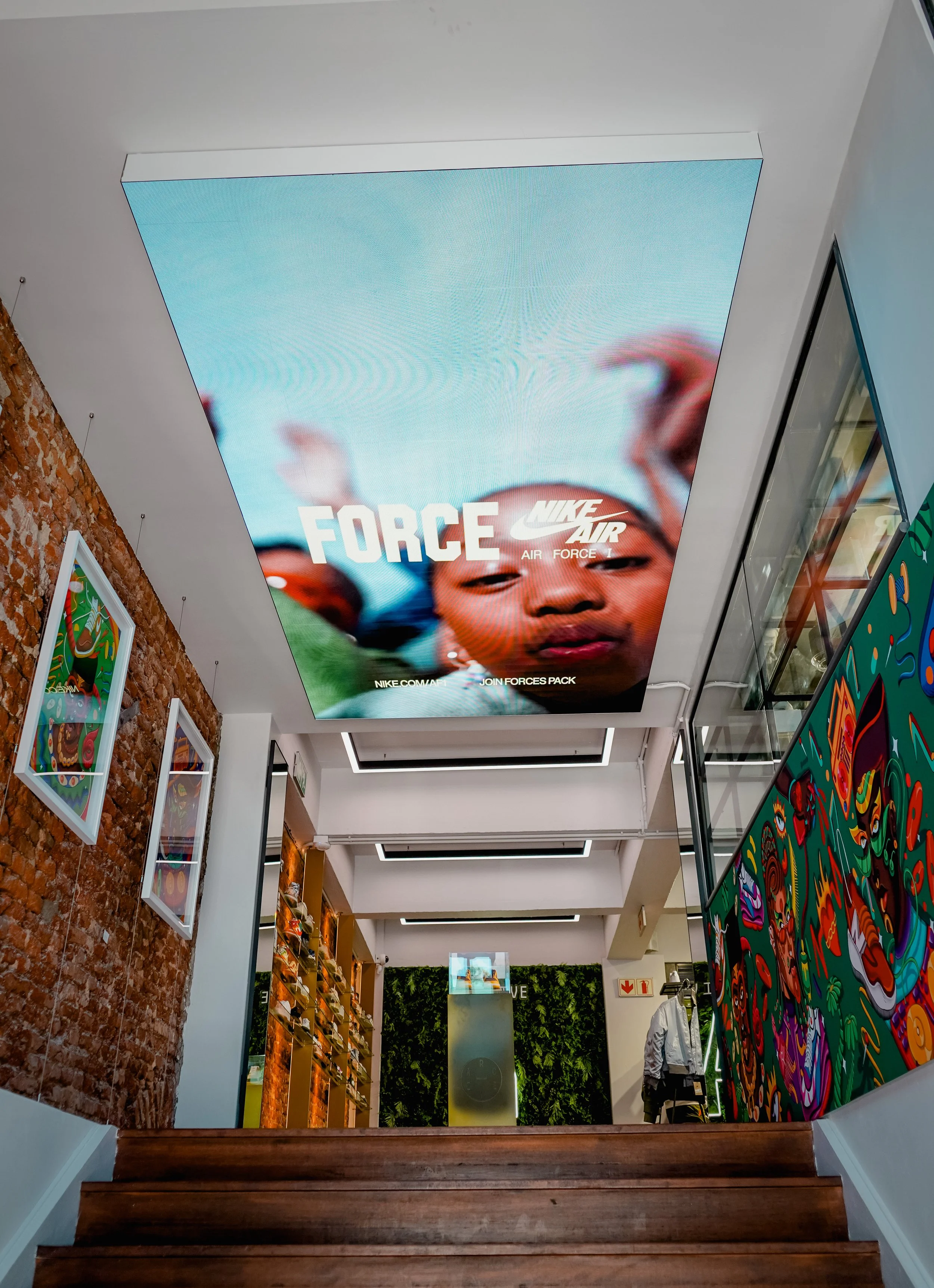 View from the bottom of a brown wooden staircase looking up at a large electronic screen on the ceiling displaying an advertisement for Nike Air sneakers with the text "FORCE" and a colorful image of a person's face.