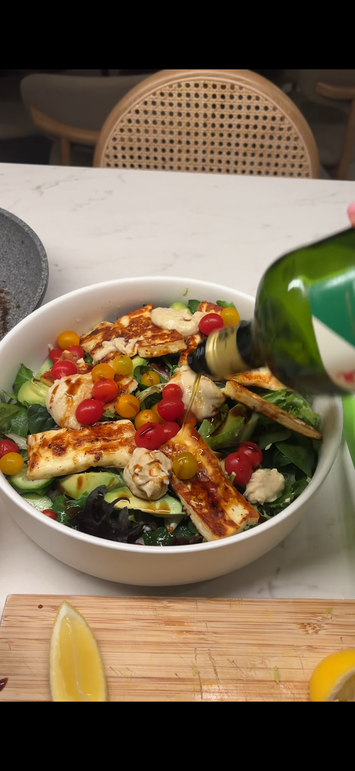 A bowl of salad with grilled chicken, cherry tomatoes, cucumbers, leafy greens, and dressing, with a bottle of dressing being poured over it.