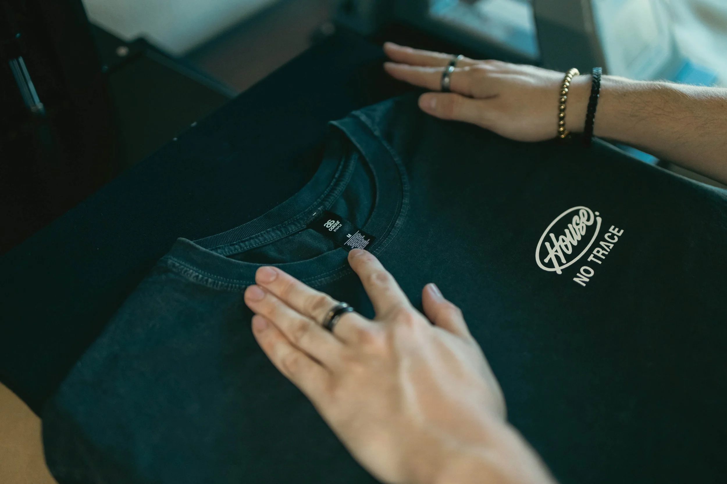 Mains posant sur un t-shirt noir avec le texte 'House No Trace' à côté d'une personne portant des bracelets et des anneaux.