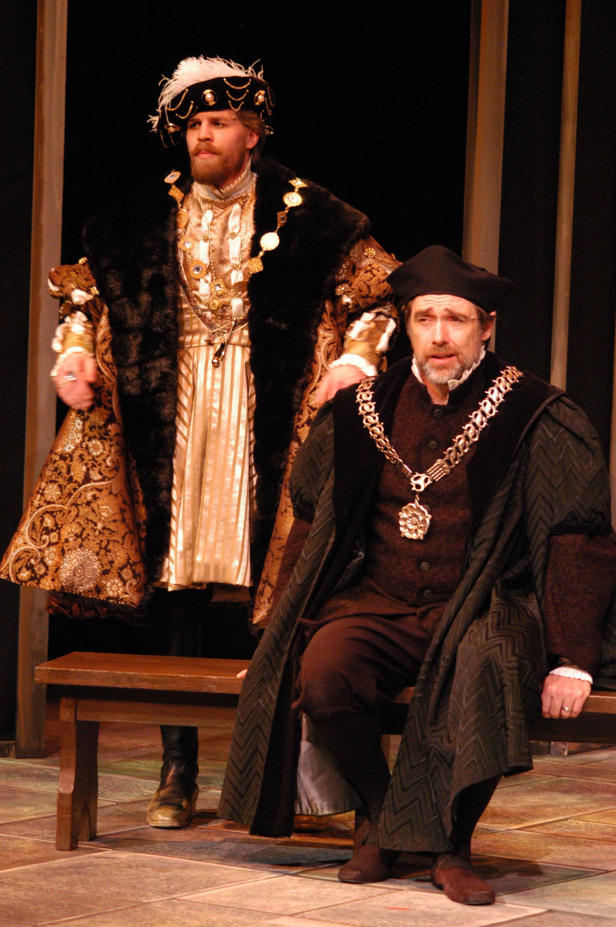 Two actors on stage dressed in elaborate historical costumes, with one standing and the other sitting, performing in a theatrical production.