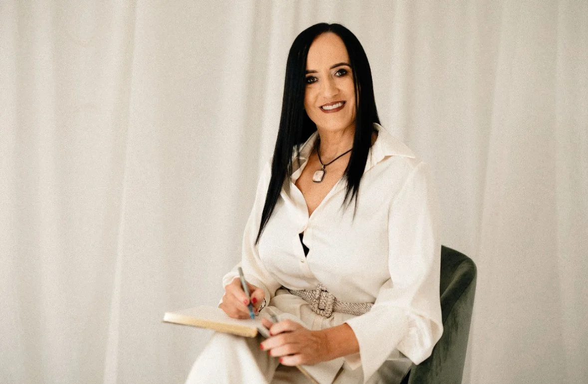 A woman with long dark hair smiling, sitting on a green chair, holding a notebook and pen, wearing a cream-colored blouse with a belt, in front of a plain light-colored background.
