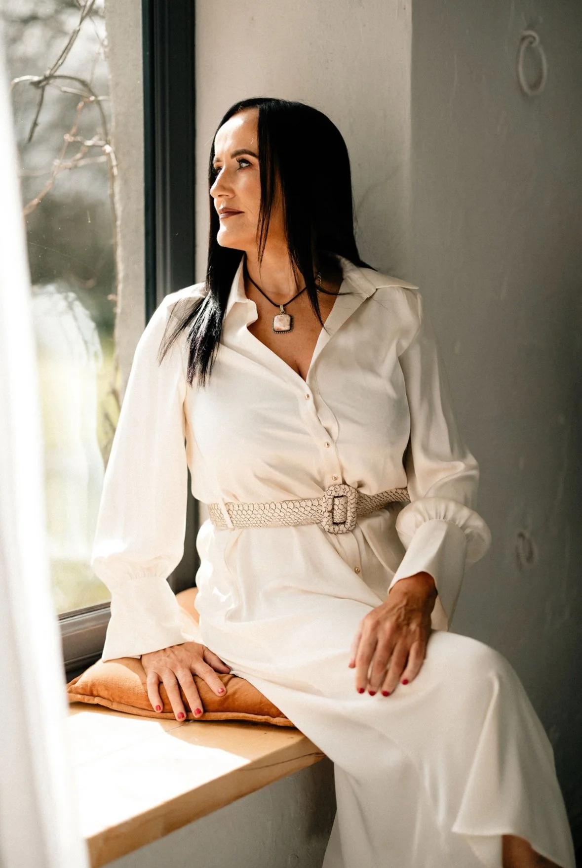A woman with black hair wearing a cream-colored dress with a belt, sitting on a windowsill with her hand resting on a pillow, gazing outside.