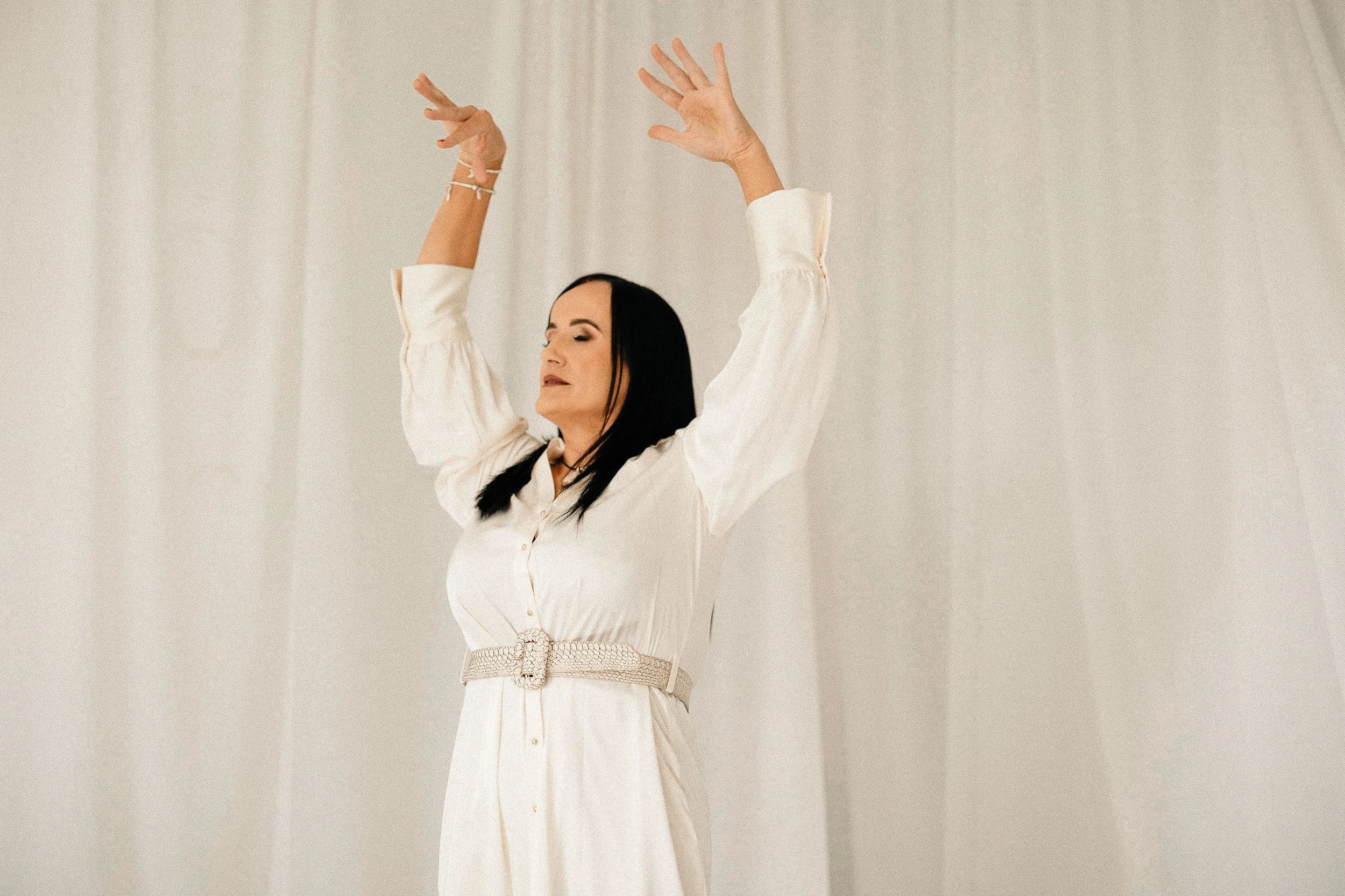 Woman dressed in an elegant white outfit with open long sleeves, raising her arms with closed eyes, standing against a neutral background.