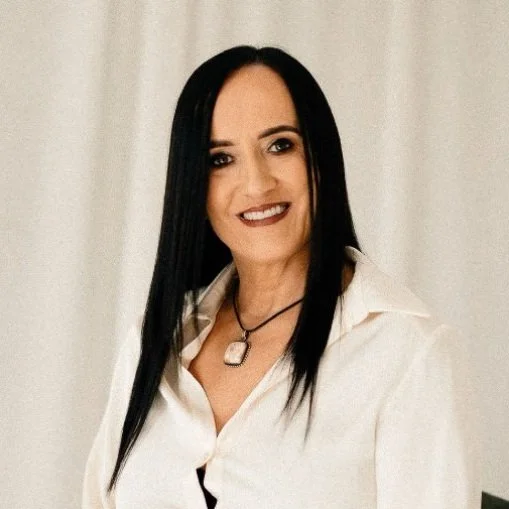 A woman with straight black hair wearing a white blouse and necklace, smiling in front of a neutral background.