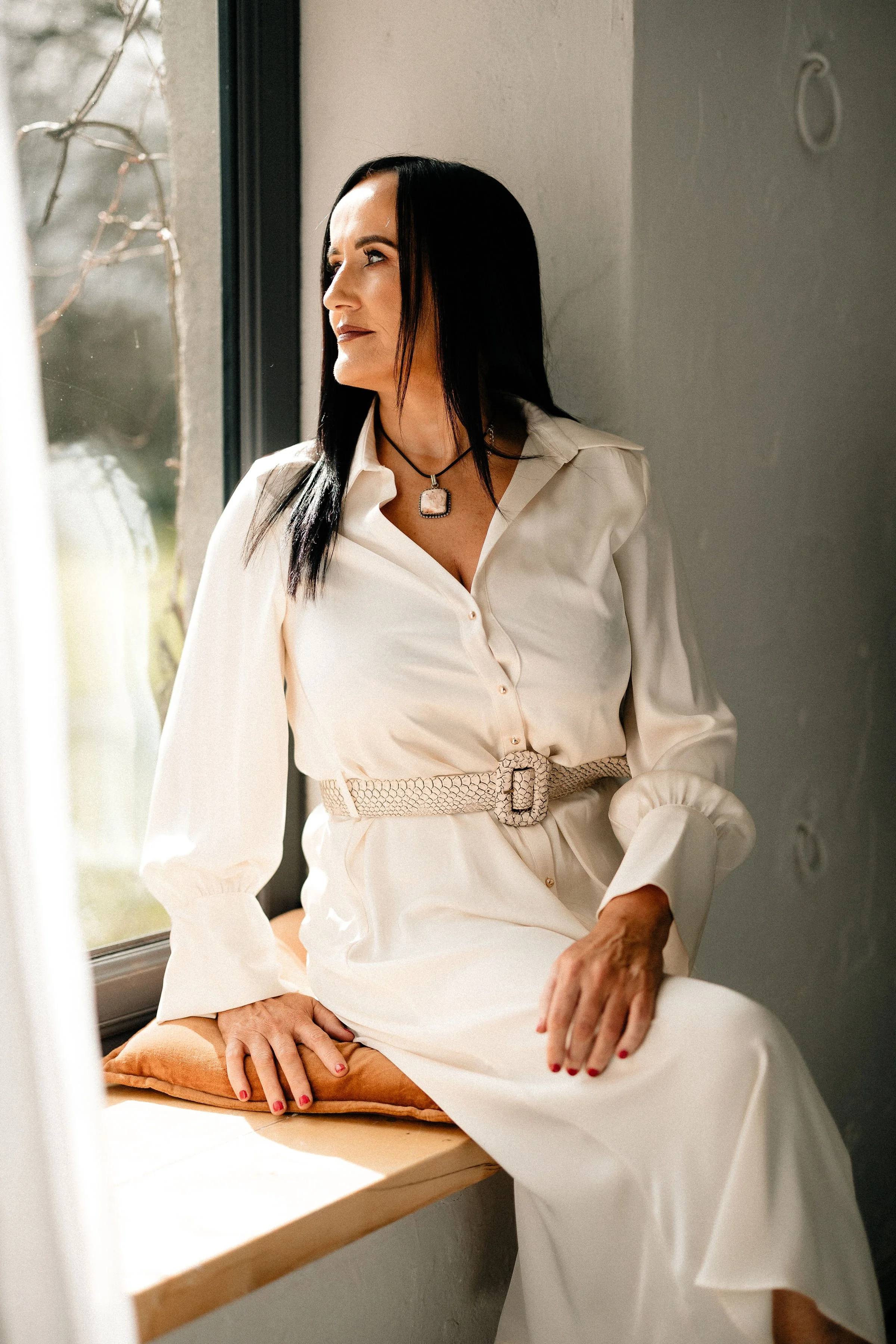 A woman sitting on a wooden window seat near a window, looking out thoughtfully, wearing a cream-colored dress with a wide belt and a square pendant necklace.