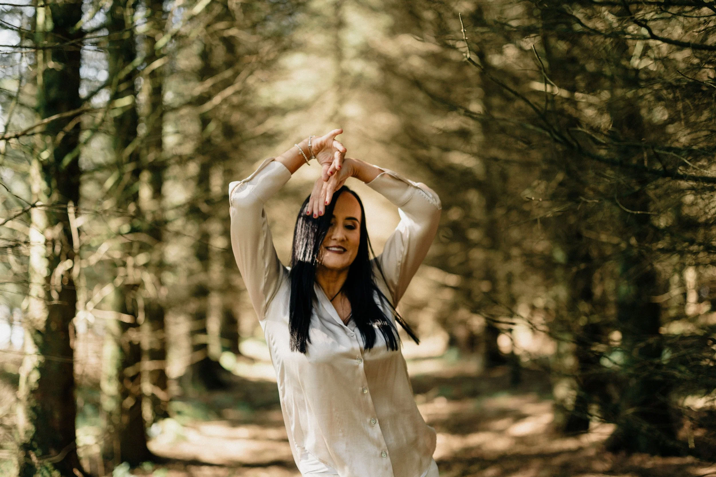 A woman with long dark hair wearing a cream-colored satin shirt standing in a sunlit forest.
