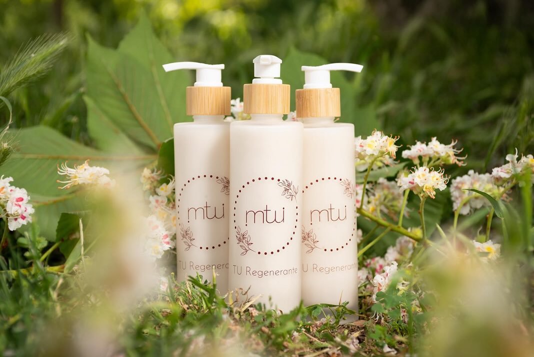 Tres botellas blancas de crema o loción con etiquetas que dicen 'mtui' y 'Tu Regenerante', colocadas en un entorno natural con flores y hojas verdes.