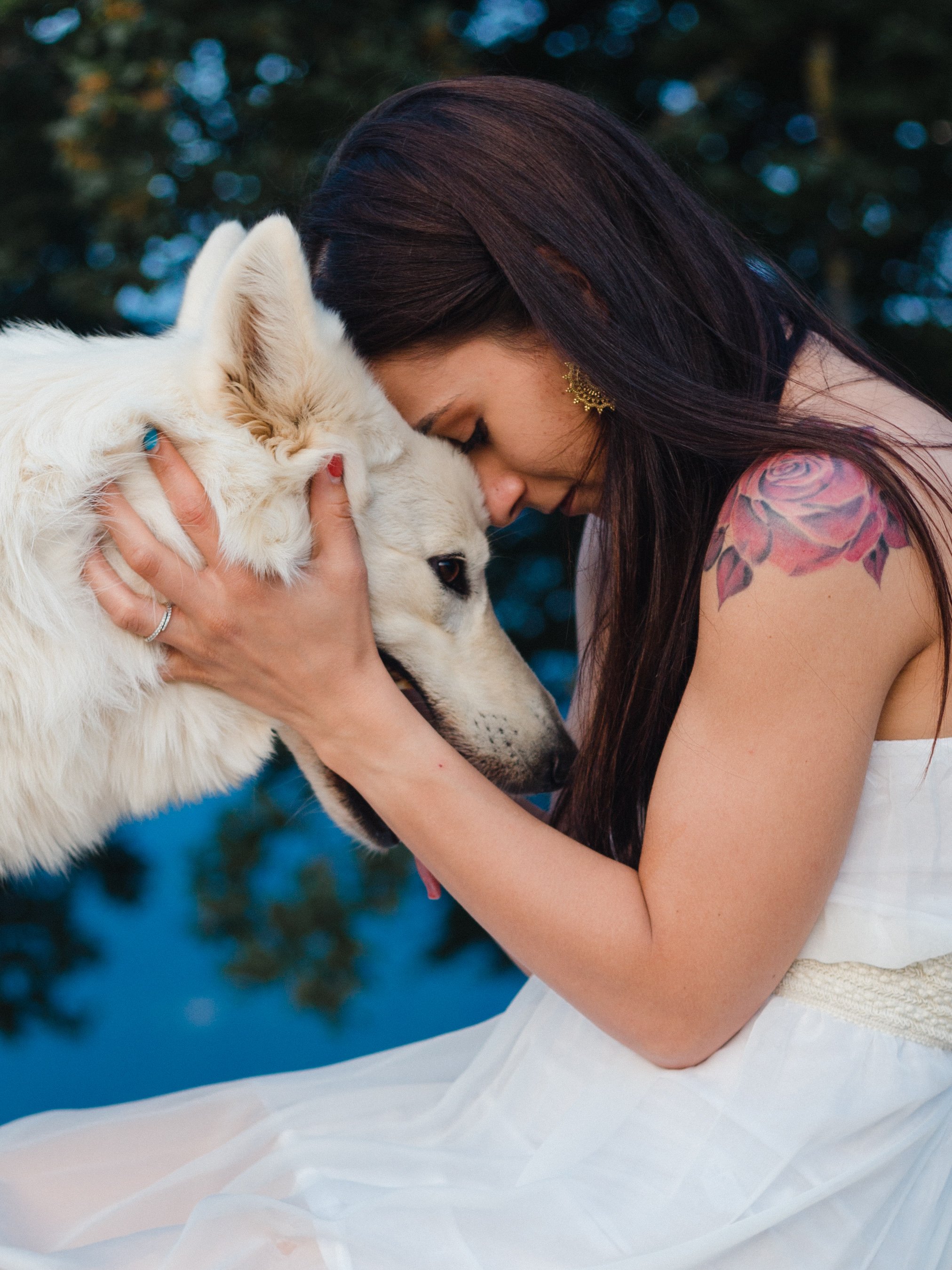 Una mujer con un tatuaje en el brazo y con cabello largo y oscuro, abrazando y tocando la cabeza de un perro blanco de raza pastor. La mujer lleva un vestido blanco y está en un entorno natural con árboles en el fondo.