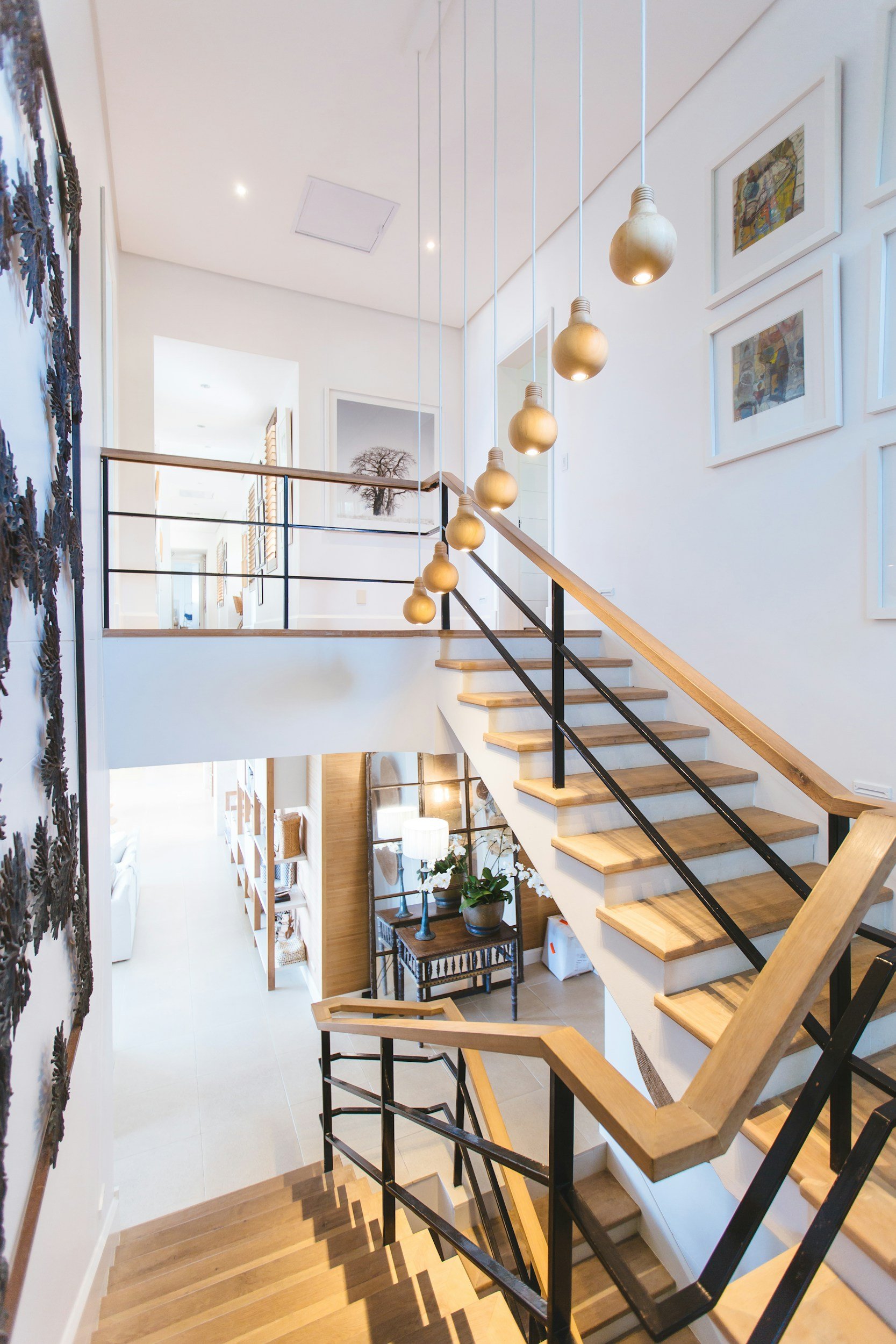 Inside view of a modern home staircase with wooden steps, black metal handrails, and hanging light fixtures shaped like light bulbs. The staircase overlooks a living room with framed artwork on the wall, a side table with plants, and a cozy seating area visible in the background. The interior has white walls and a high ceiling with recessed lighting.