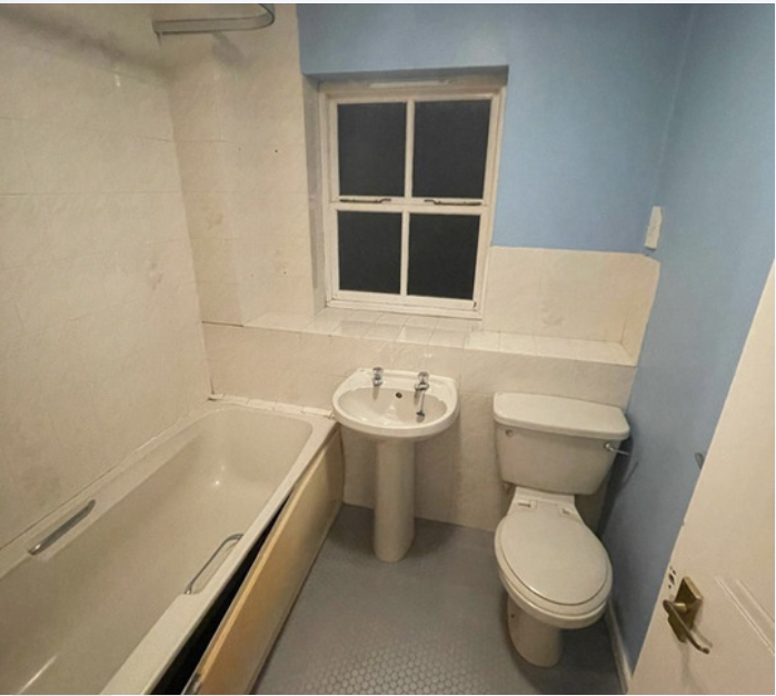 Small bathroom with a bathtub, a pedestal sink, and a toilet, with a window above the sink, painted light blue and beige walls, and a white door.