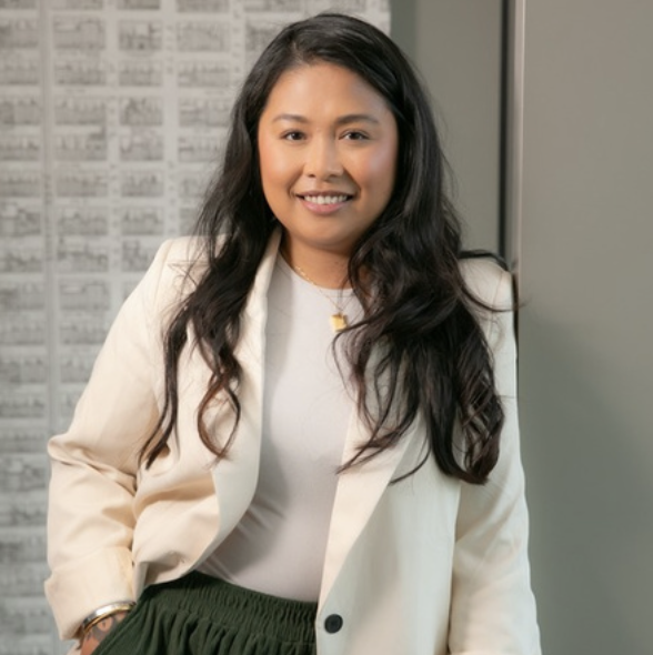A smiling woman with long dark hair, wearing a white blazer over a white top, standing indoors with a gray wall and a pattern of numbers or letters behind her.