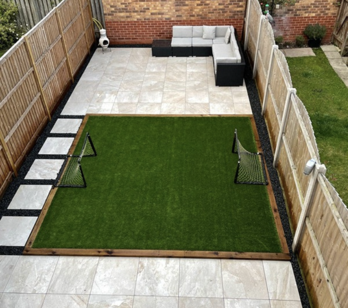 Backyard with a green artificial grass area, two small soccer goals, stone pathway, patio with outdoor sofa, and wooden fencing.