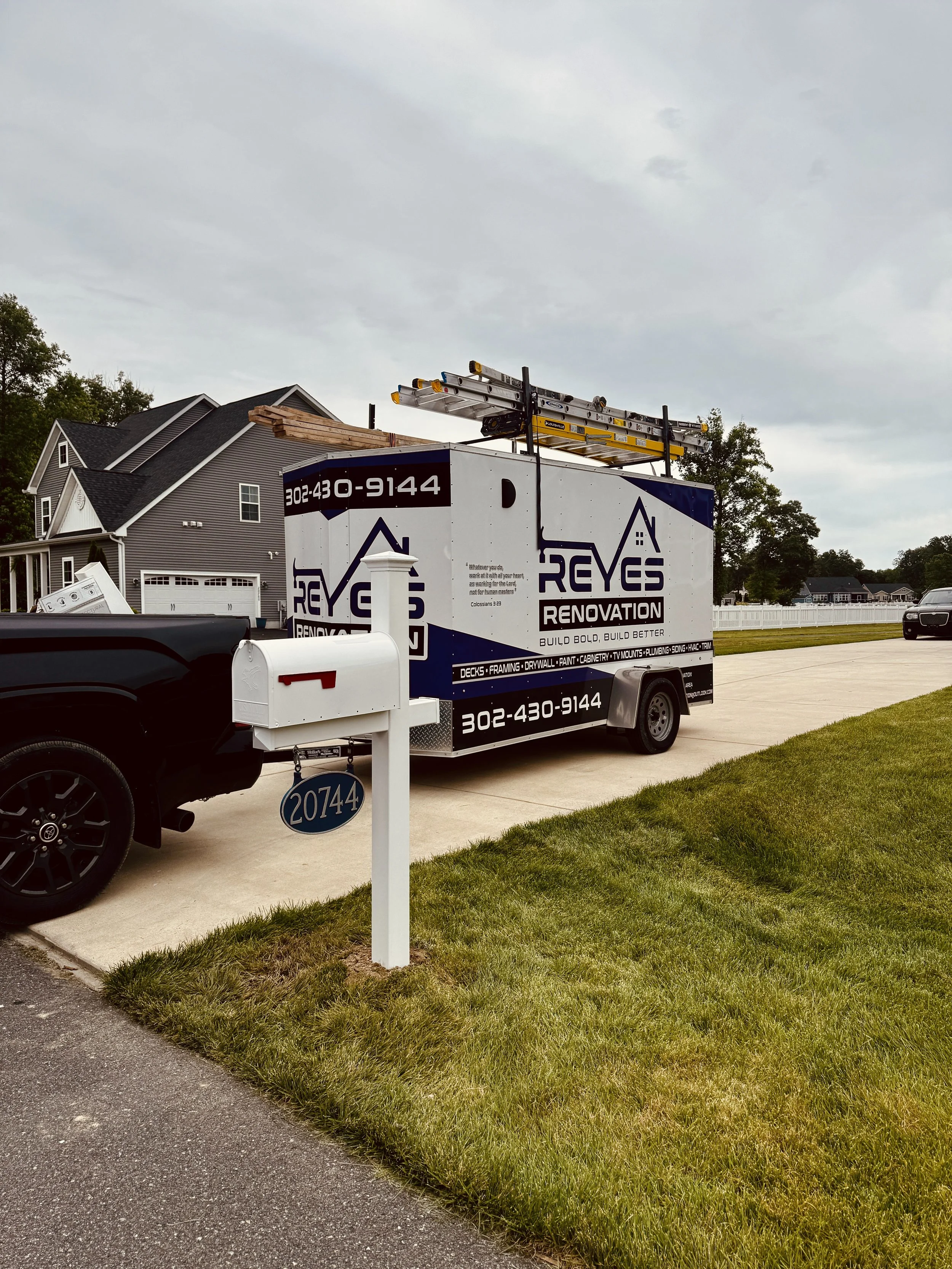A black pickup truck parked next to a white mailbox with a yard address sign reading 20744, and a black and white trailer with roofing company branding in a suburban neighborhood, with houses and trees in the background.