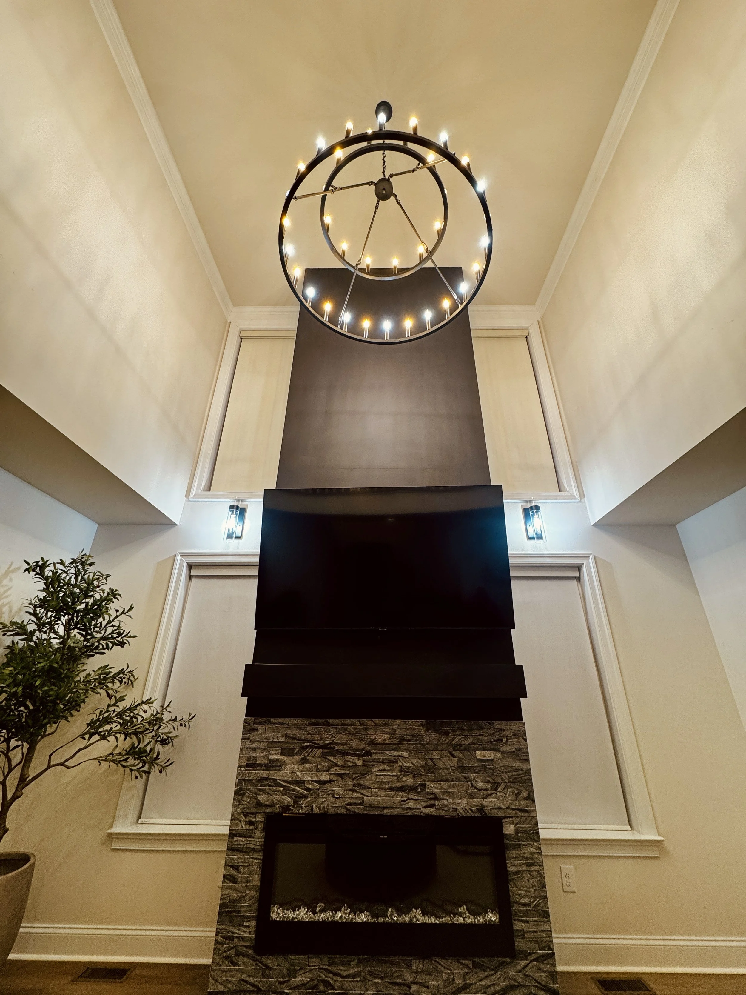 Living room with a high ceiling, modern fireplace with stacked stone surround, large TV, wall sconces, and a chandelier resembling a clock with lights.