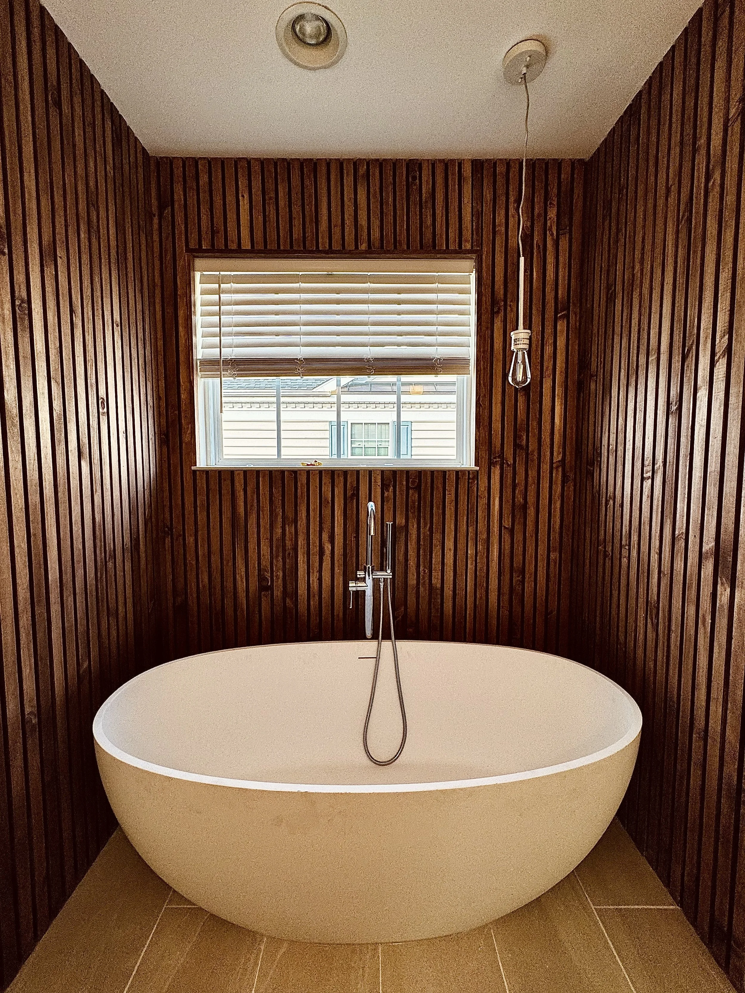 A bathroom with wood-paneled walls, a freestanding oval bathtub, a window with blinds, and a hanging light fixture.