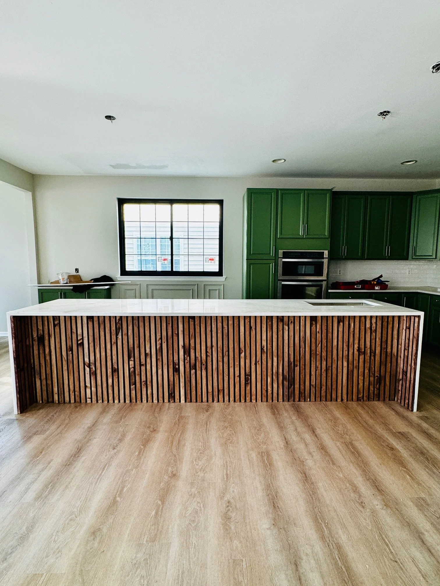Modern kitchen with green cabinets, a white countertop, and a wood-paneled island, with a window on the back wall and a light-colored wood floor.