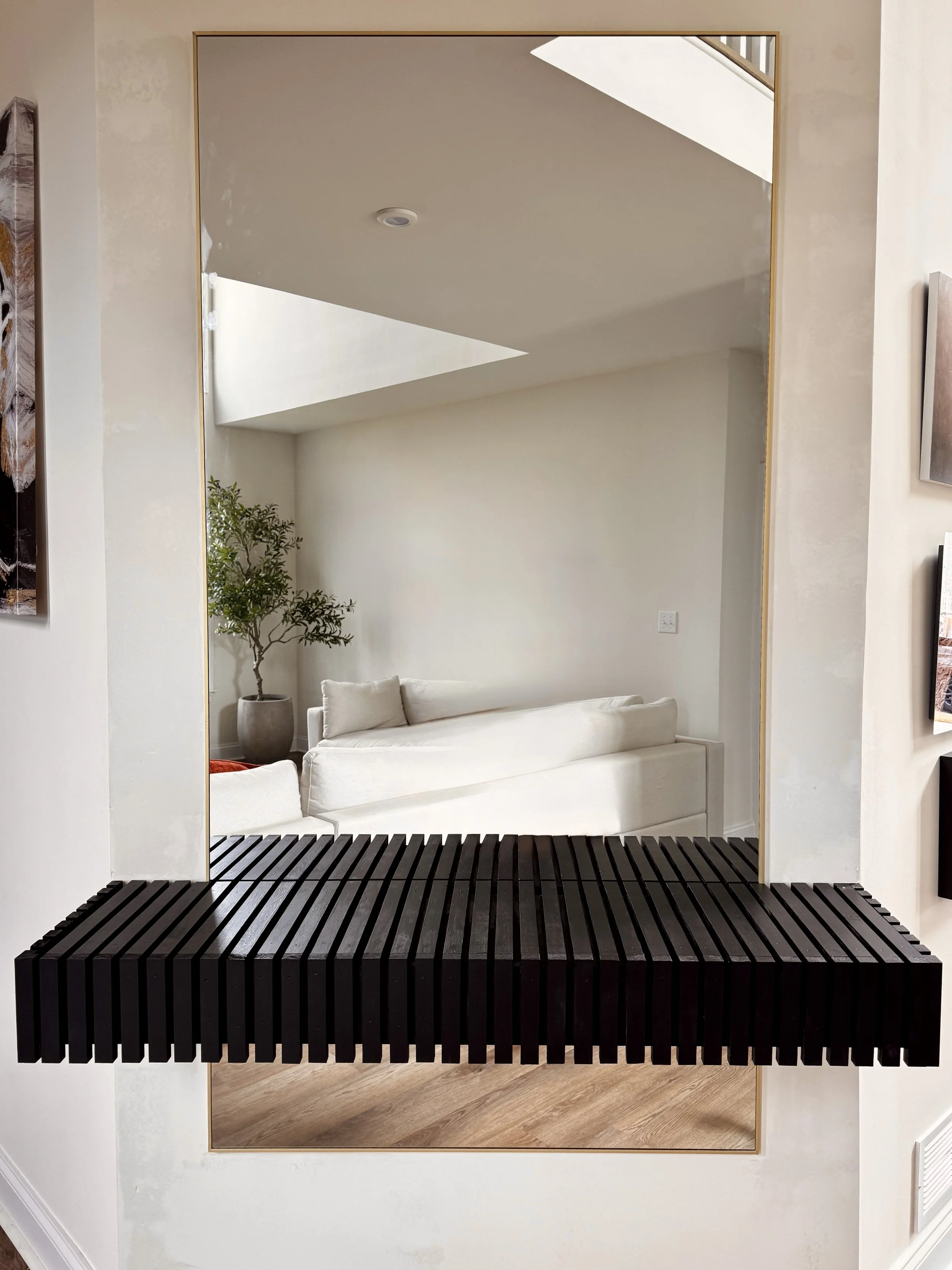 A large rectangular mirror with a white wall backdrop. In front of the mirror, there is a black wooden slatted bench or shelf extending from the wall. The reflection shows a white couch with a small pillow and a potted plant with green foliage. The ceiling has a skylight allowing natural light to illuminate the room.