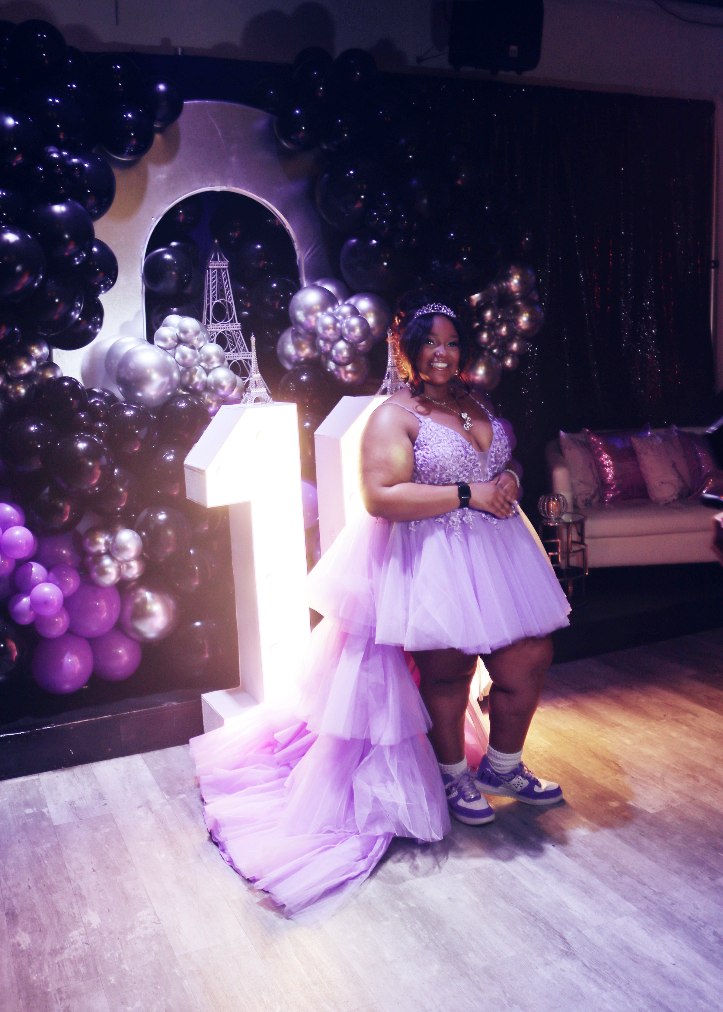 A woman in a lavender dress with a voluminous tulle skirt stands in front of a birthday party backdrop decorated with black, purple, and metallic balloons, illuminated number '13' signs, and a metallic Eiffel Tower replica.