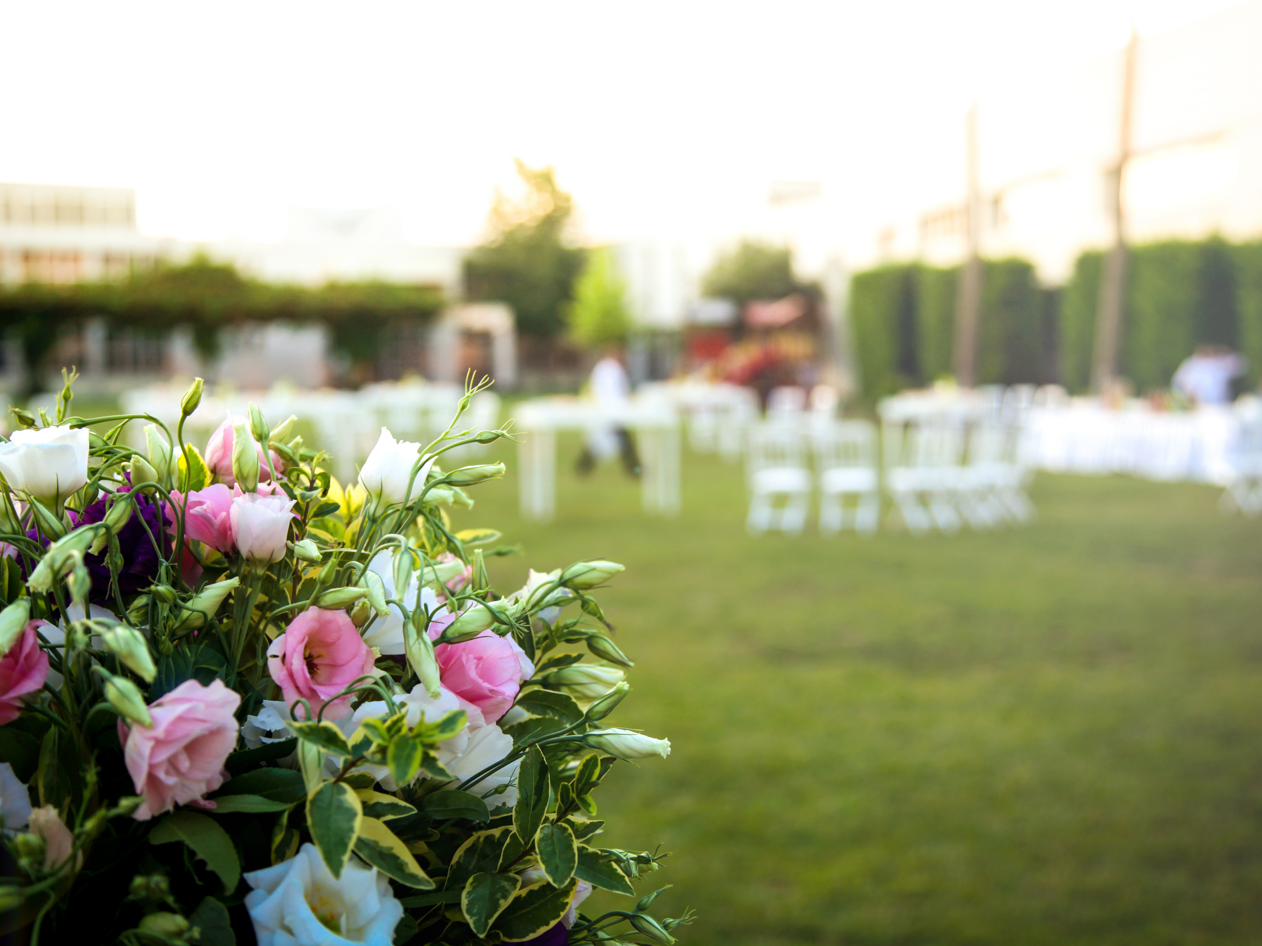 Close-up of pink, white, and purple flowers in the foreground at an outdoor event with blurred chairs and people in the background.