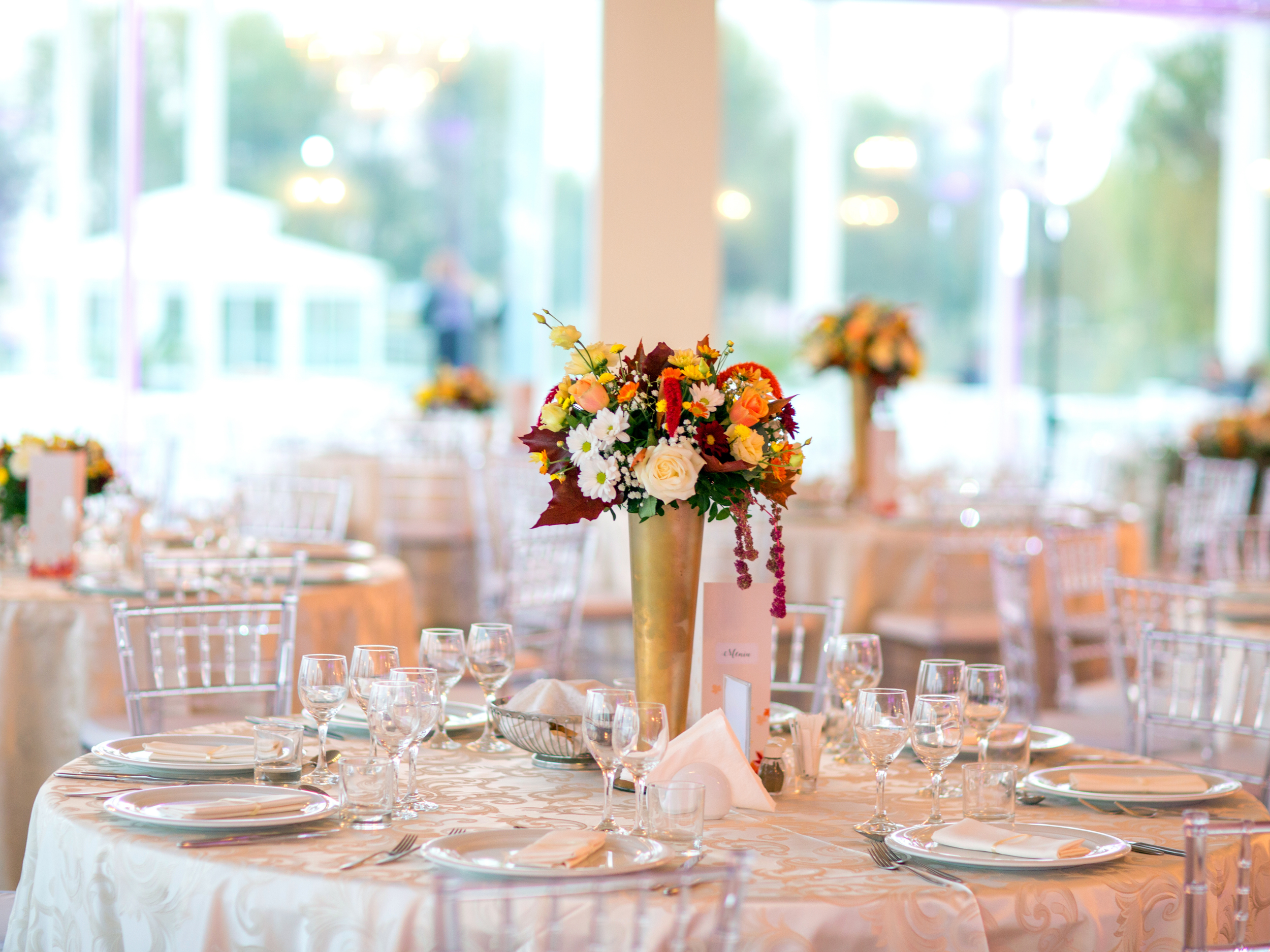 Elegant event setup with round tables, white tablecloths, clear chairs, and central floral arrangements in gold vases featuring roses, daisies, and colorful flowers, set with plates, glasses, and napkins in a bright, modern venue with large windows.