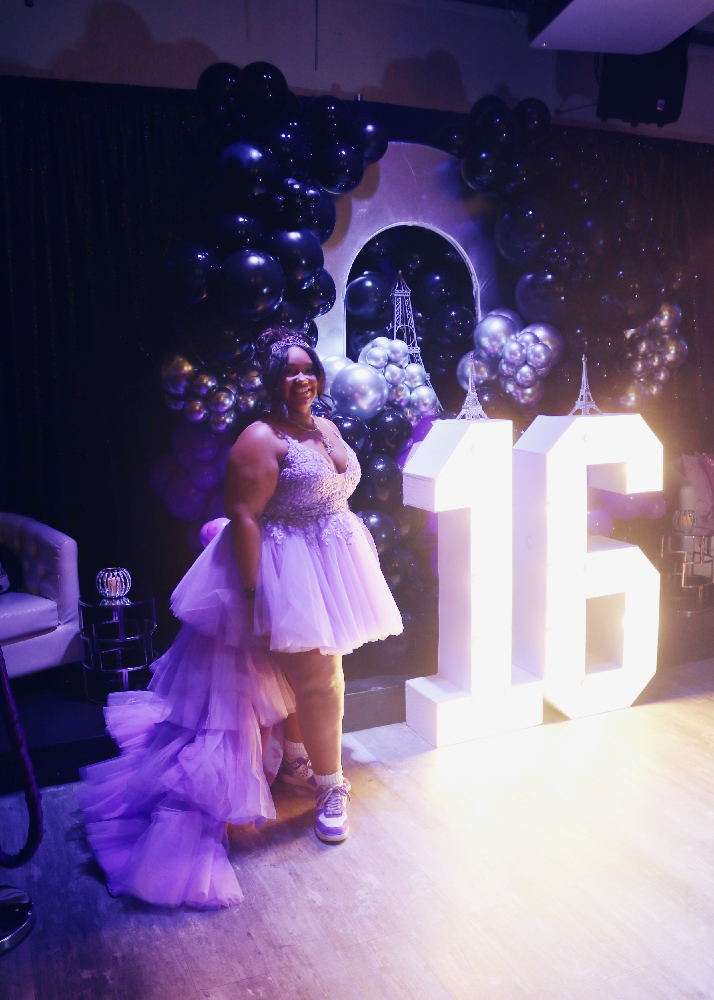 A woman in a purple ball gown standing next to large illuminated number 15 and Eiffel Tower decorations for a milestone birthday celebration.