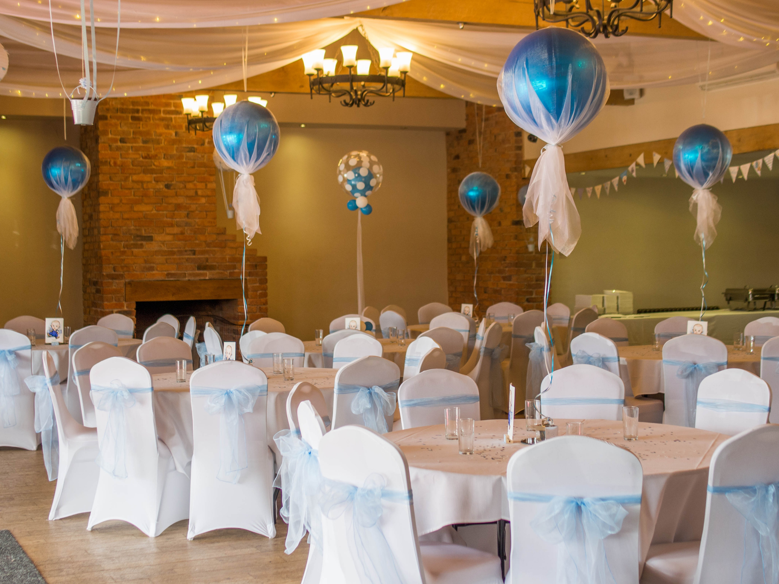 A decorated banquet hall with round tables, white chairs with light blue sashes, blue balloons tied with white tissue paper, a brick fireplace, and hanging white fabric with string lights.