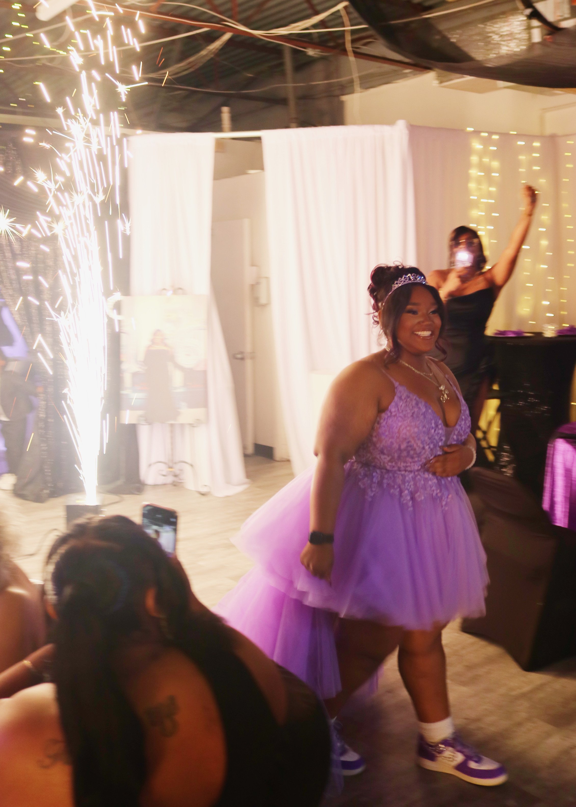 Celebration with a woman in a purple dress and tiara smiling, surrounded by sparklers and friends, at an indoor party with fairy lights.