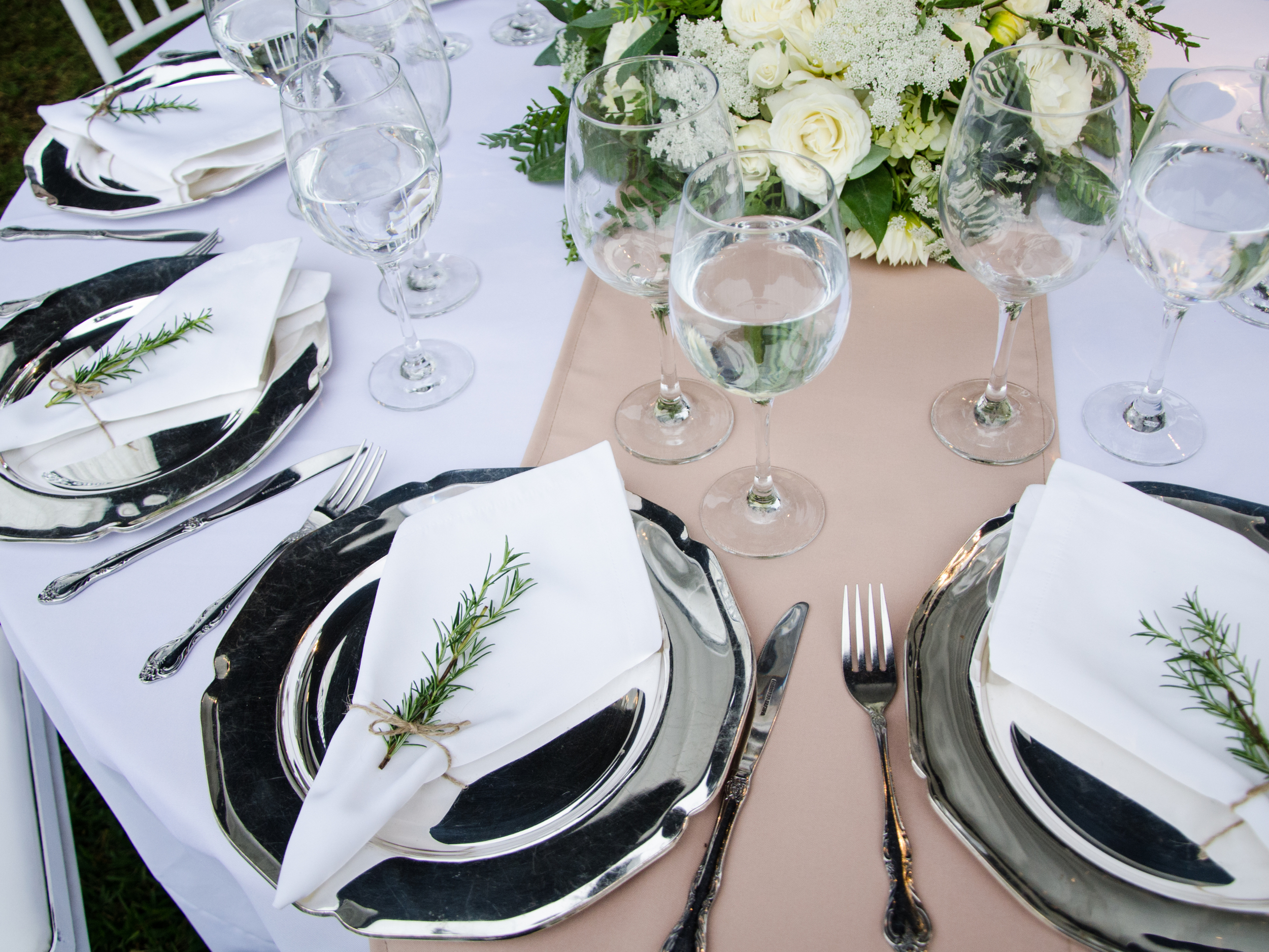 Elegant table setting with silver chargers, white napkins with sprigs of rosemary, multiple wine glasses, a floral centerpiece with white roses and greenery, and a table runner.