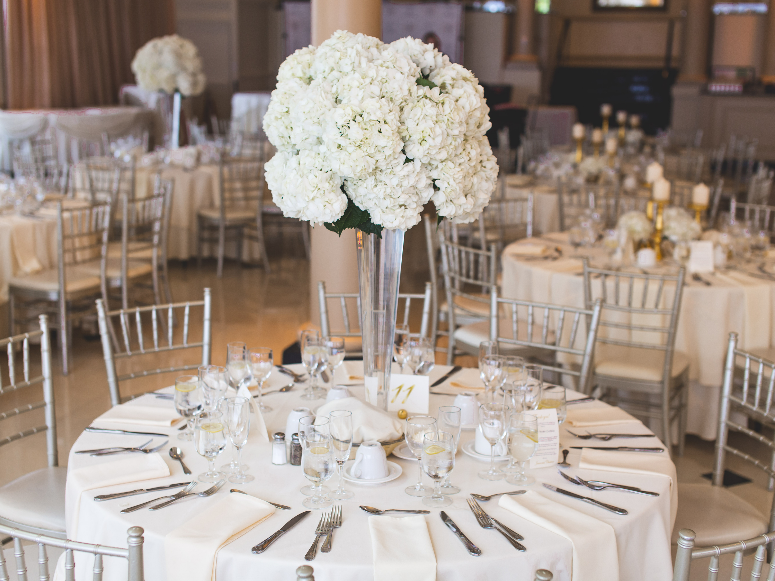Elegant banquet table with white tablecloth, silver chairs, and a tall floral centerpiece of white hydrangeas in a glass vase at a formal event