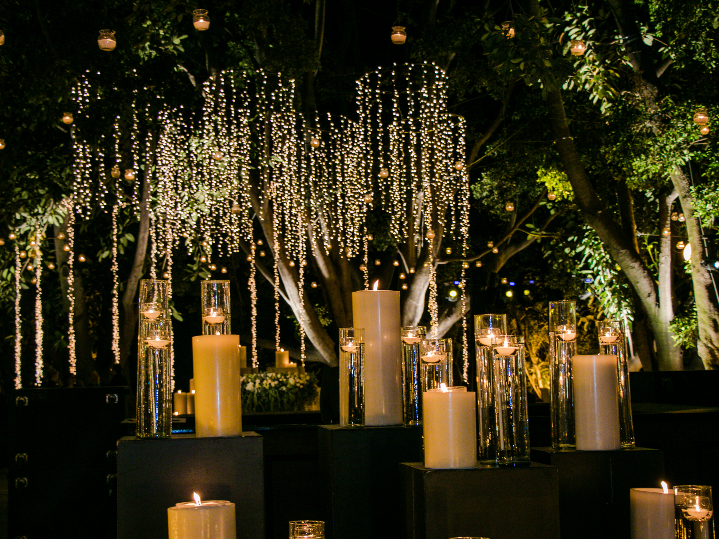 Decorative outdoor scene with illuminated hanging string lights, tall candles in glass holders, and lush green trees in the background.