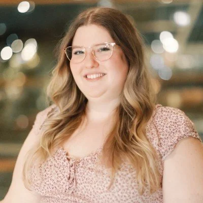 A woman with glasses and long, wavy hair smiling in front of a blurred background with lights.