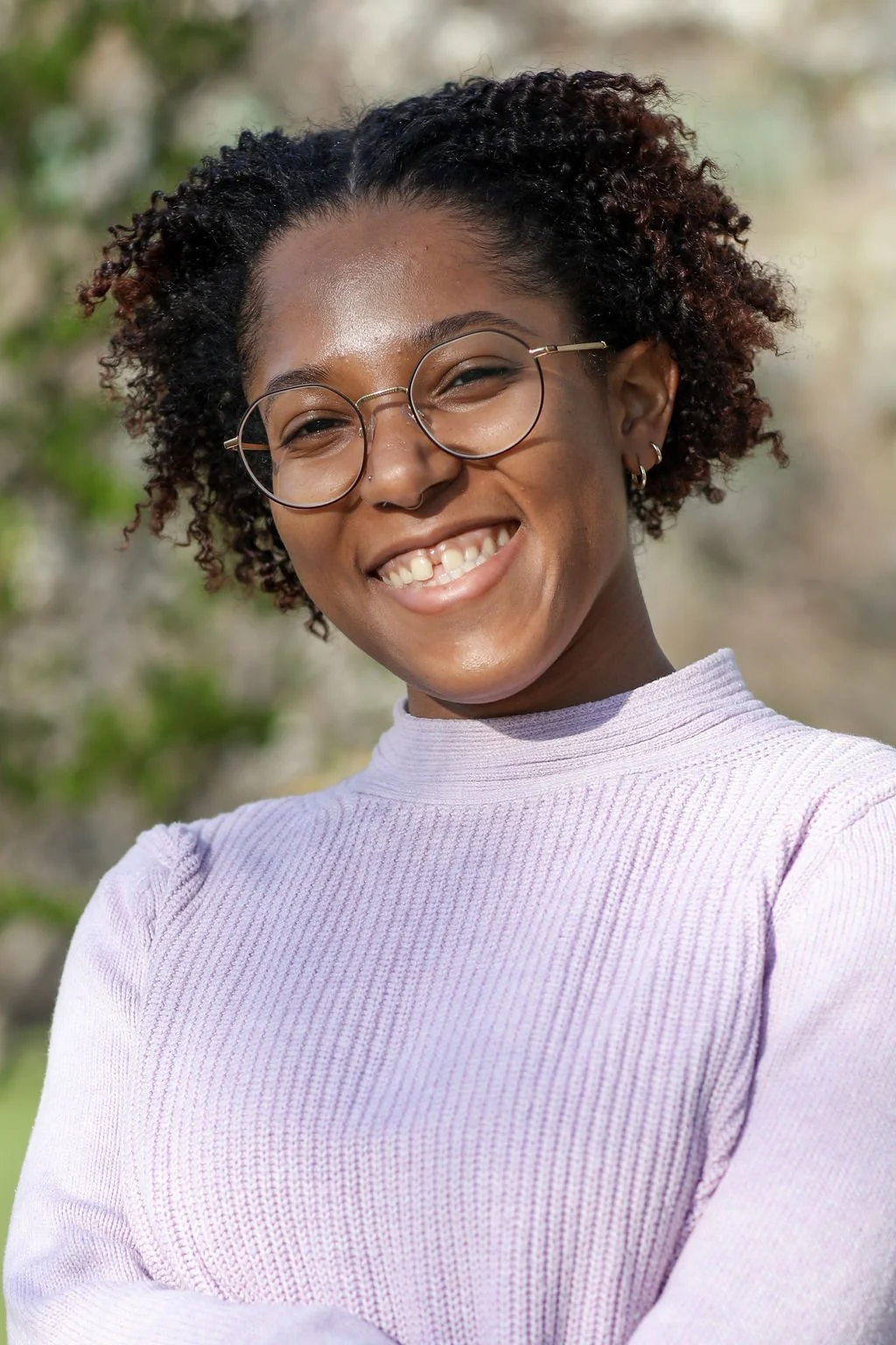A young Black woman with curly hair and round glasses smiling outdoors in natural light.