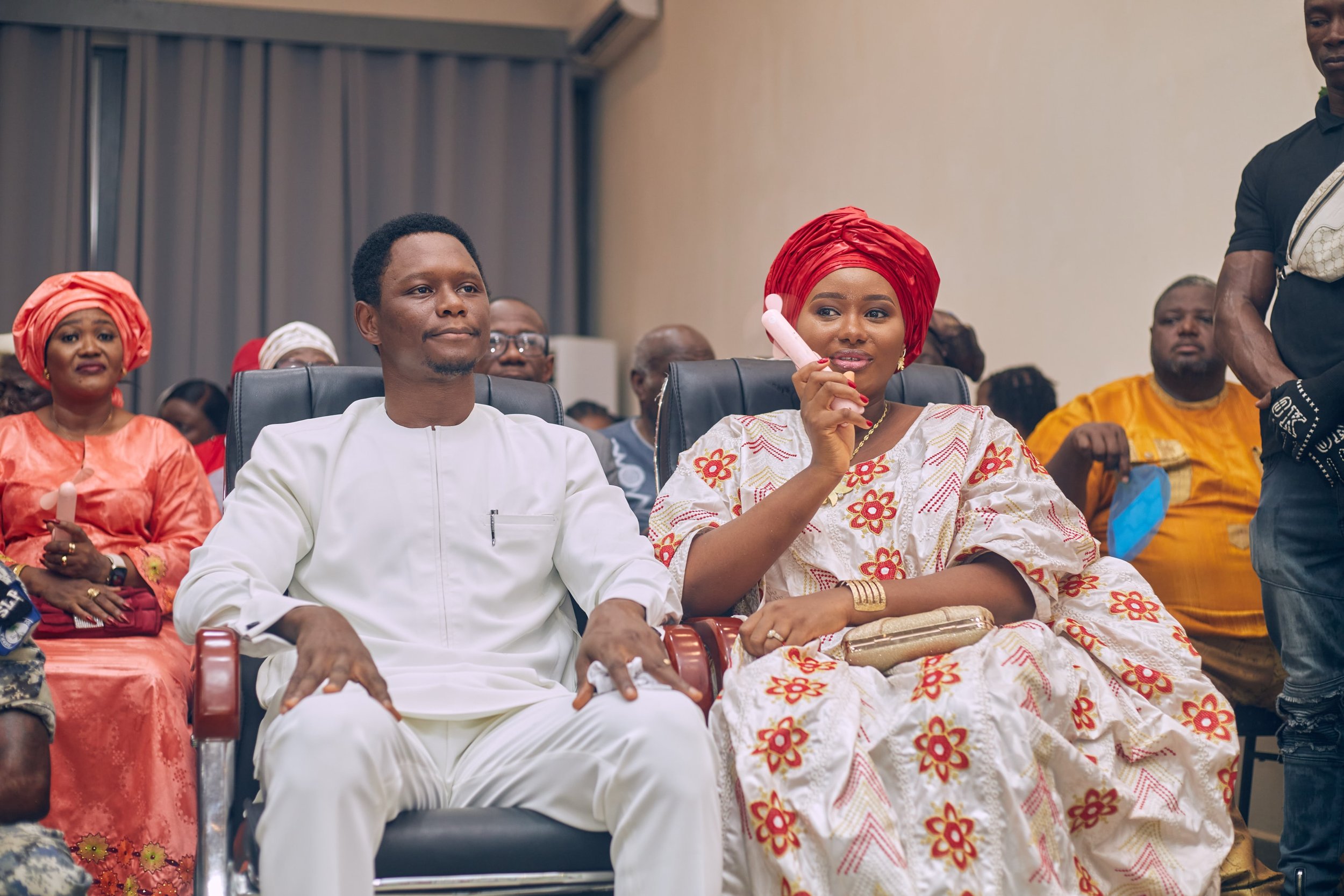 A group of people attending a cultural event, with a woman in colorful traditional attire and head wrap sitting next to a man in a white outfit, and other attendees in the background.