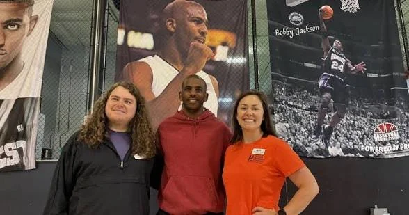 Three people standing in front of large posters of basketball players, one of whom is Chris Paul, in a sports facility or gym.