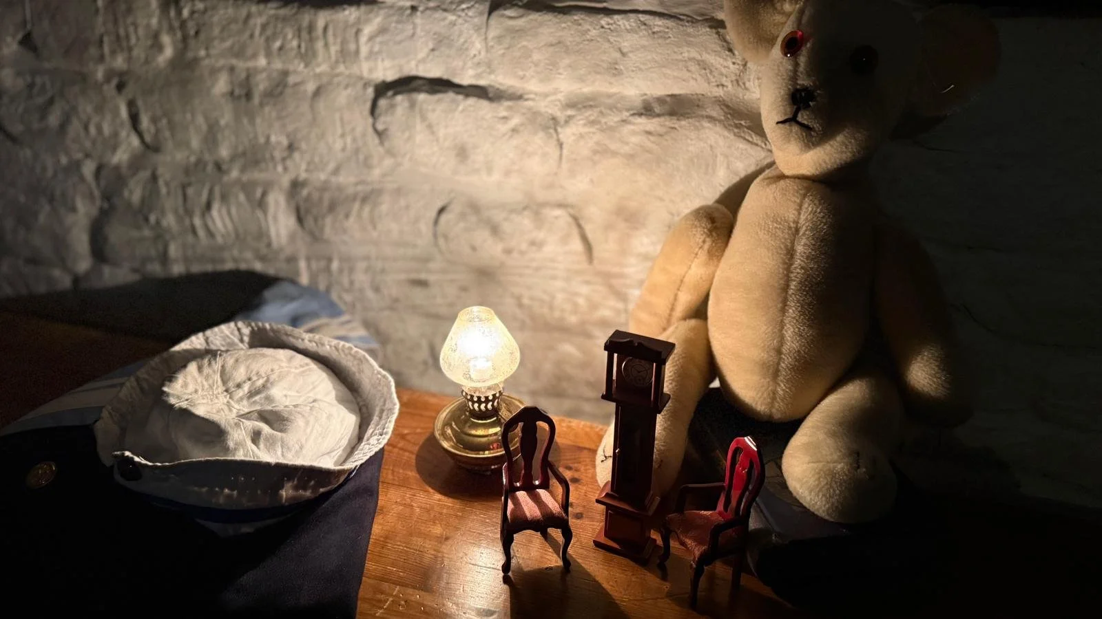 A teddy with some dolls house furniture dimly lit by a single light bulb