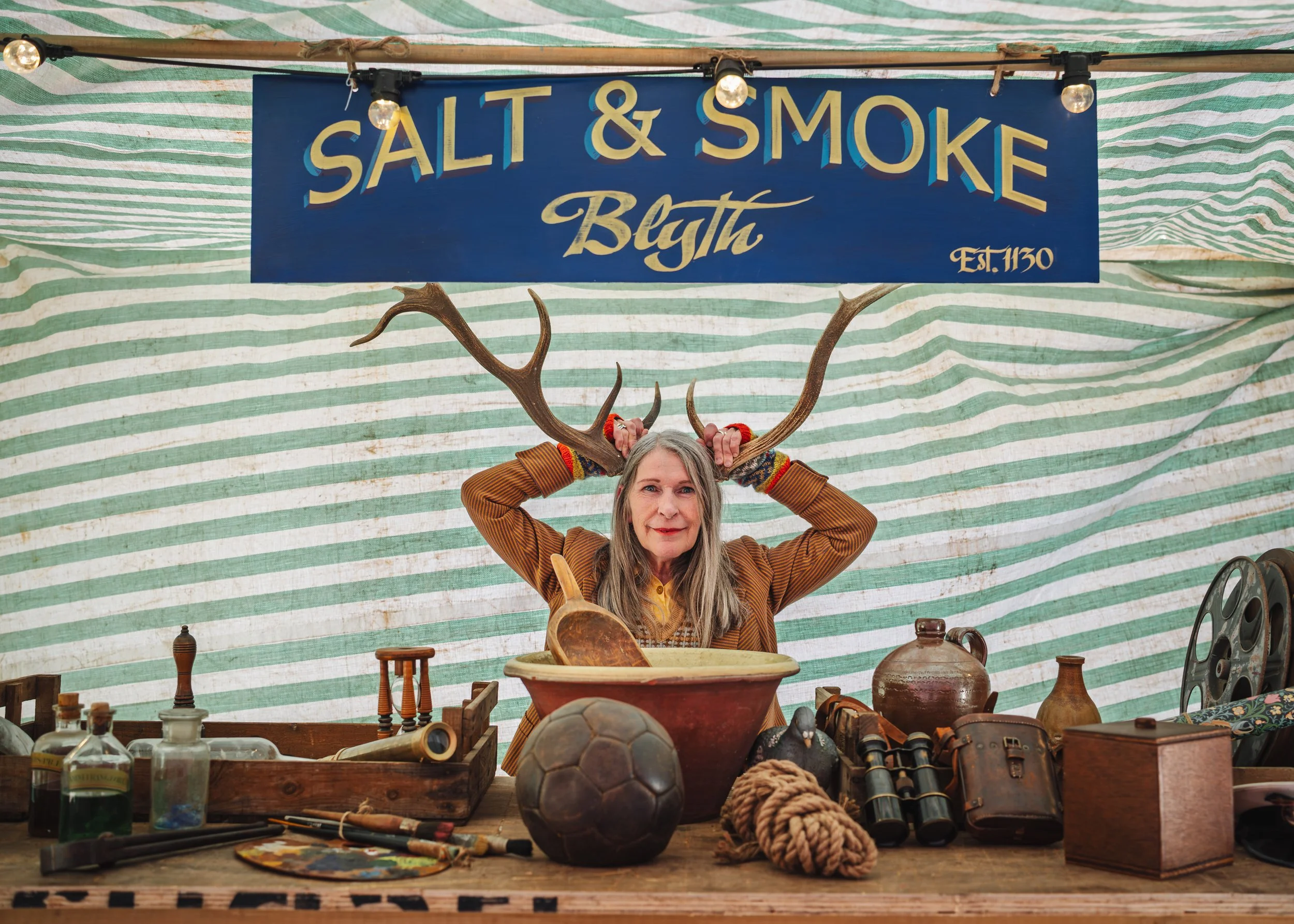 Actor holding two antlers on her head. A sign in the background says Salt and Smoke Blyth