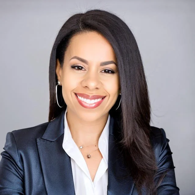 Portrait of a smiling woman with straight black hair, wearing hoop earrings, a white blouse, and a dark blazer against a plain gray background.