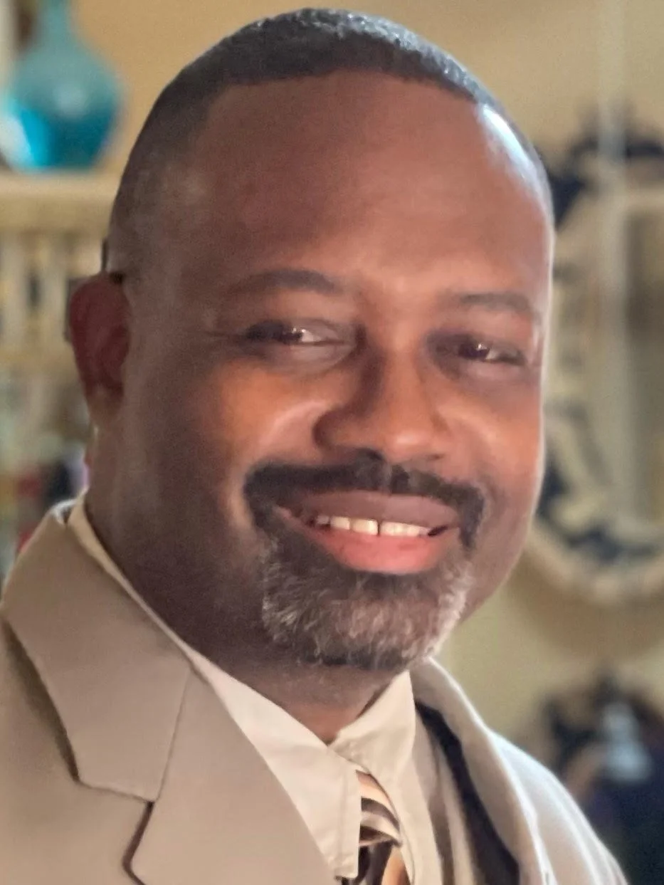 Close-up of a smiling man wearing a beige suit and a striped tie, indoors with blurred background.